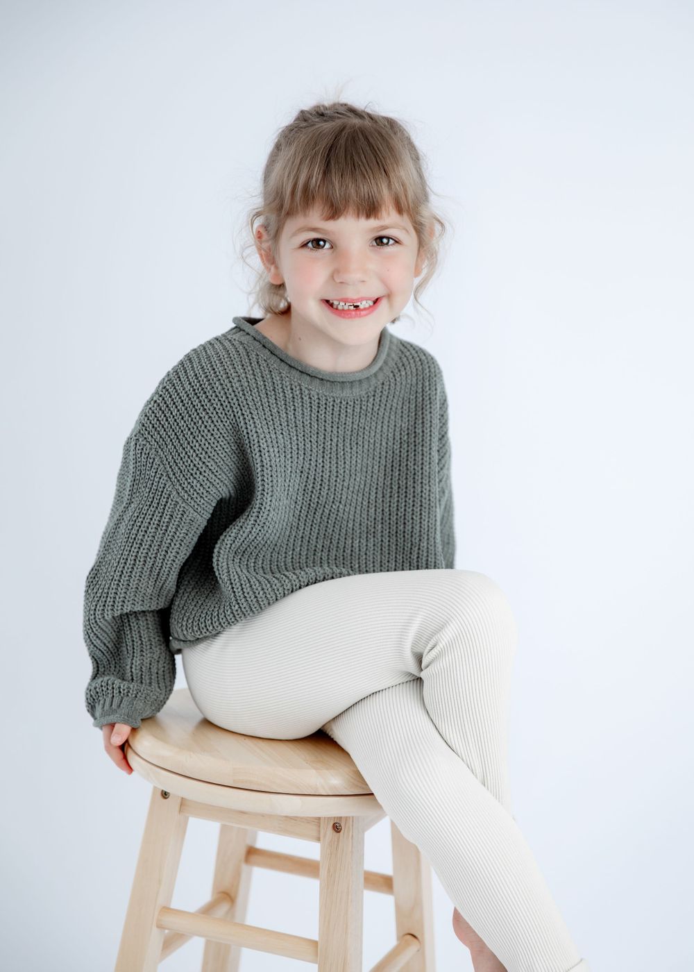 Studio shot on wooden stool wearing cozy winter sweater and cream colored pants.