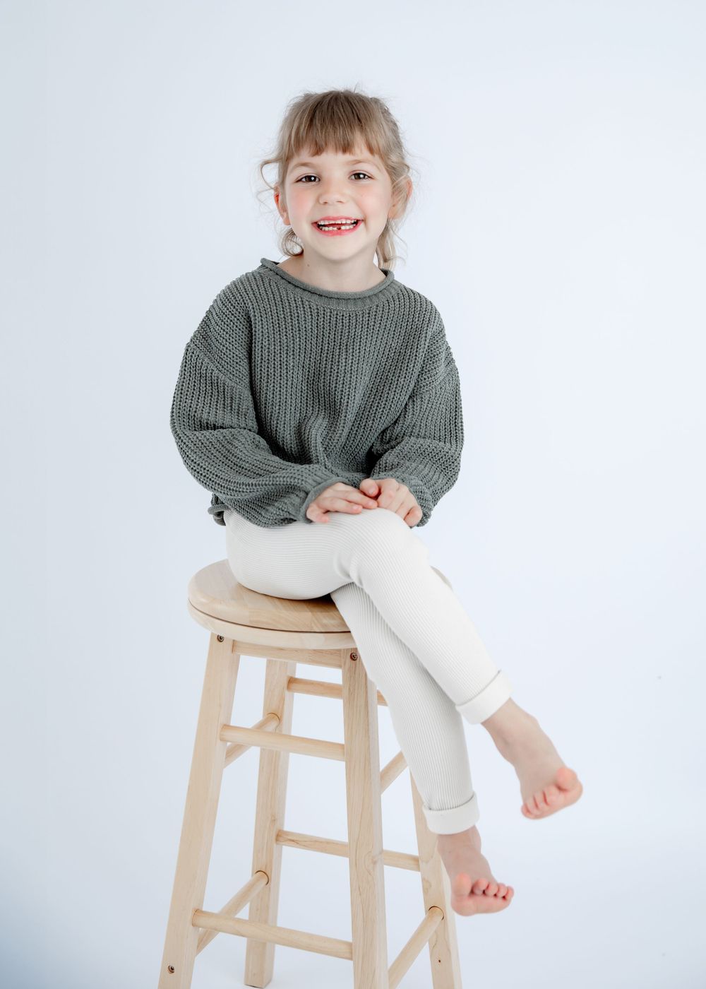 Relaxed pose on tall wooden stool wearing chunky knit sweater in minimalist setting.