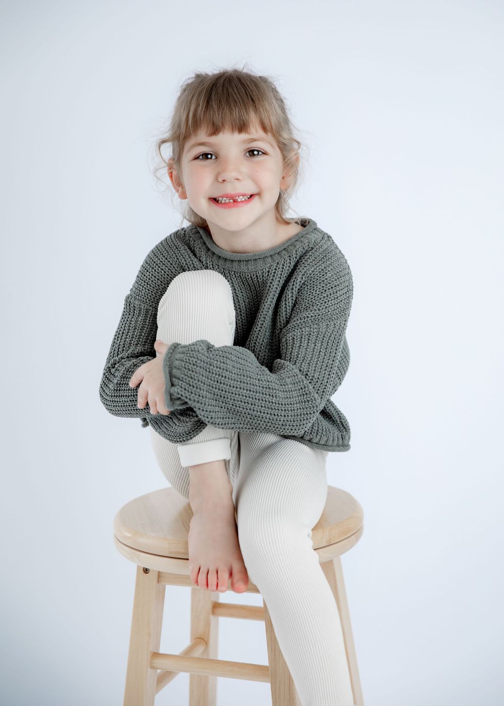 Playful studio portrait in comfortable gray sweater and light colored pants on wooden stool.