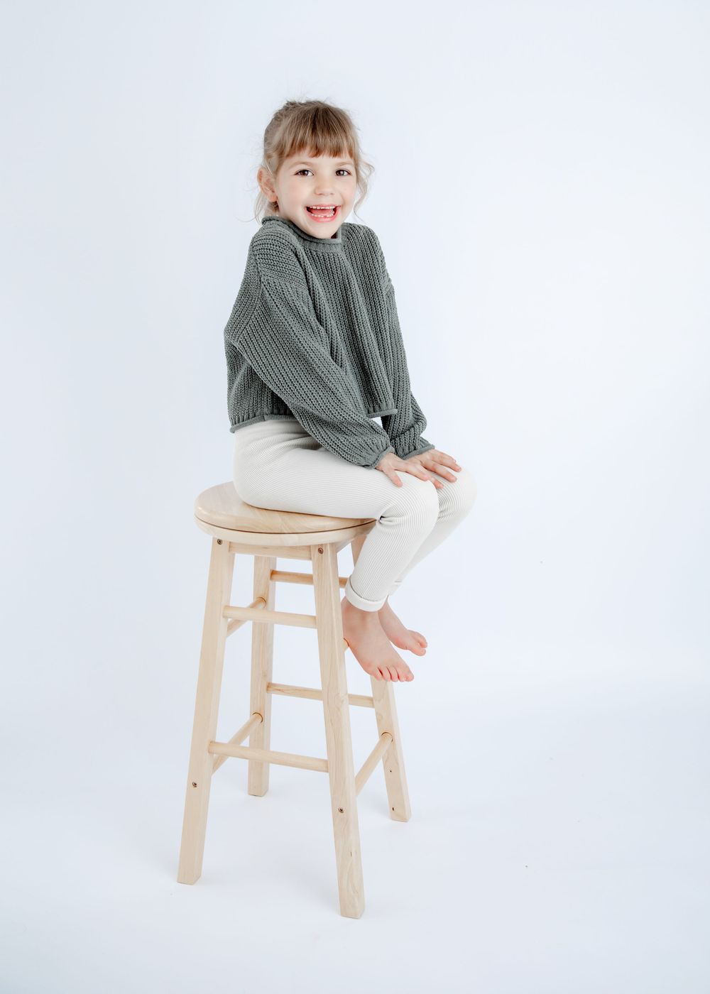 Cheerful pose on wooden barstool wearing gray sweater and white pants against white backdrop.