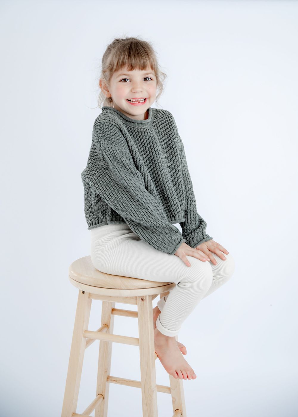 Sitting casually on wooden stool wearing gray sweater and cream pants in studio setting.
