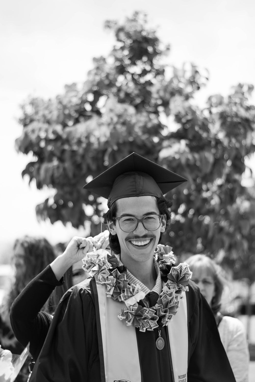 Graduate in cap and gown wearing lei and glasses smiles during commencement ceremony.