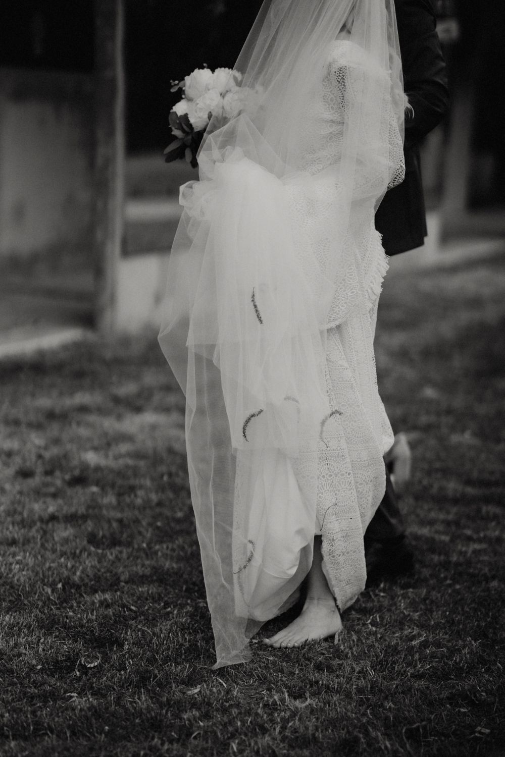 Close-up view of flowing white wedding dress and formal shoes on grass.