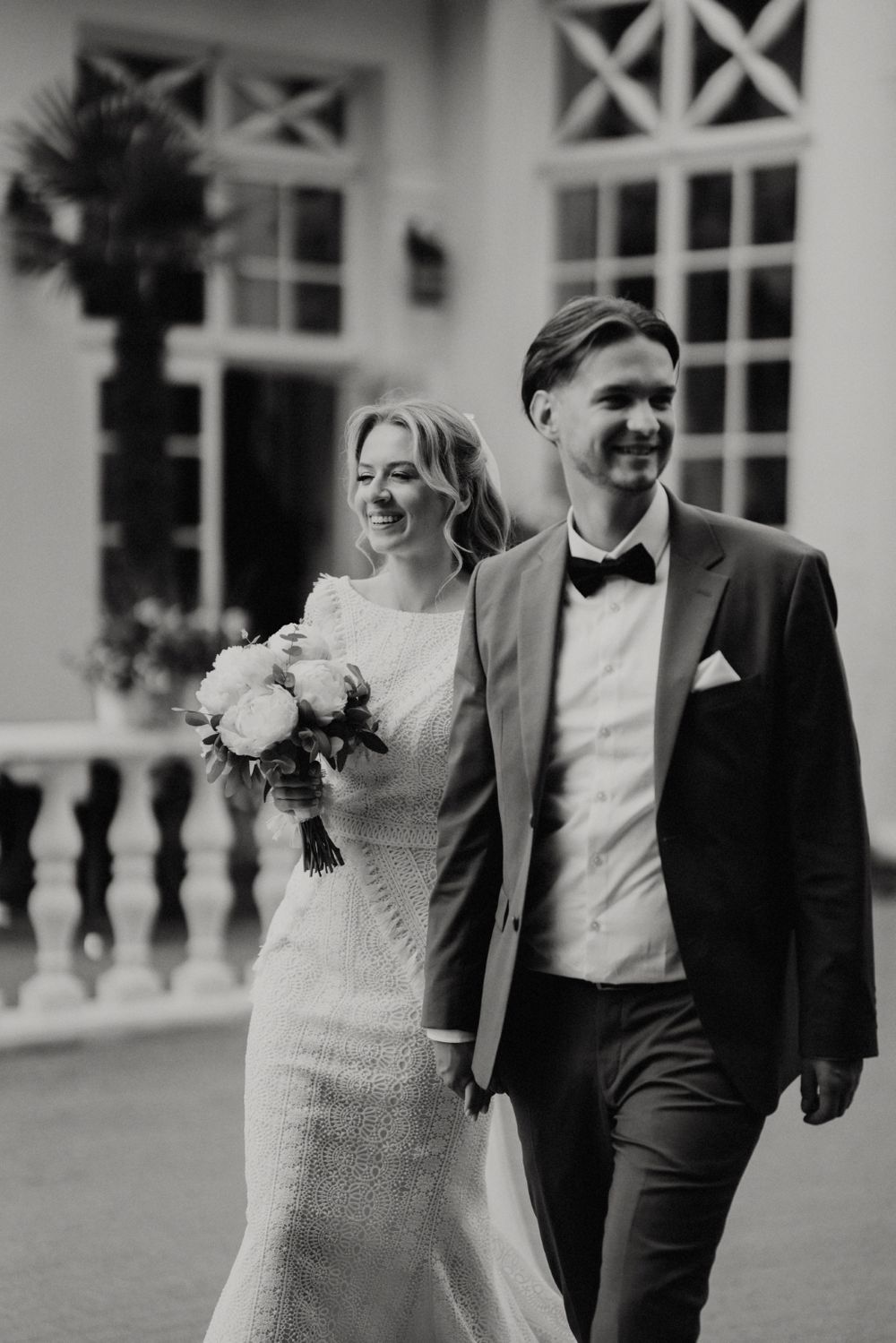 Newlyweds walk together along an elegant white building with classical architectural details.