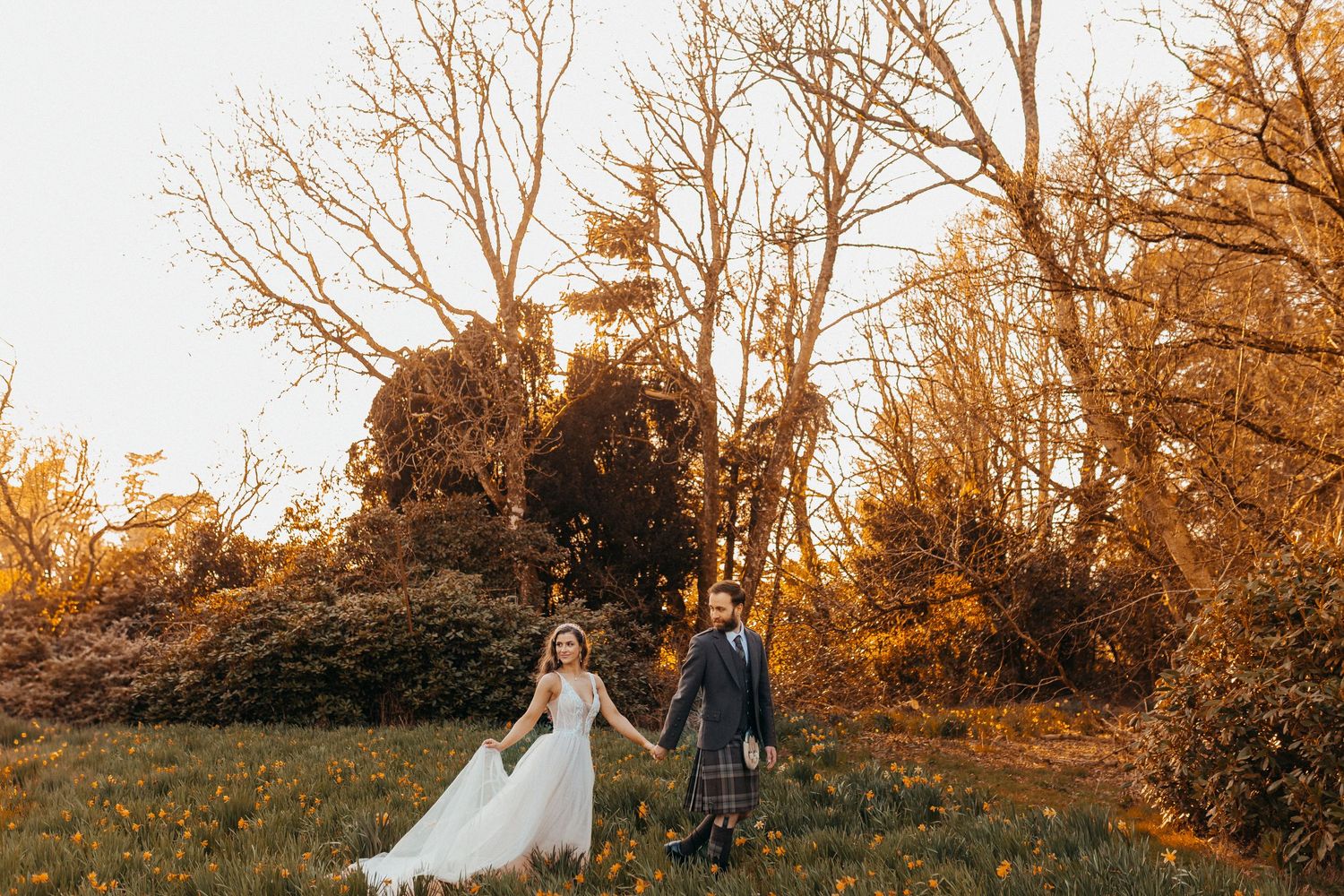 A couple in wedding attire holds hands while walking through a golden autumn field at sunset.