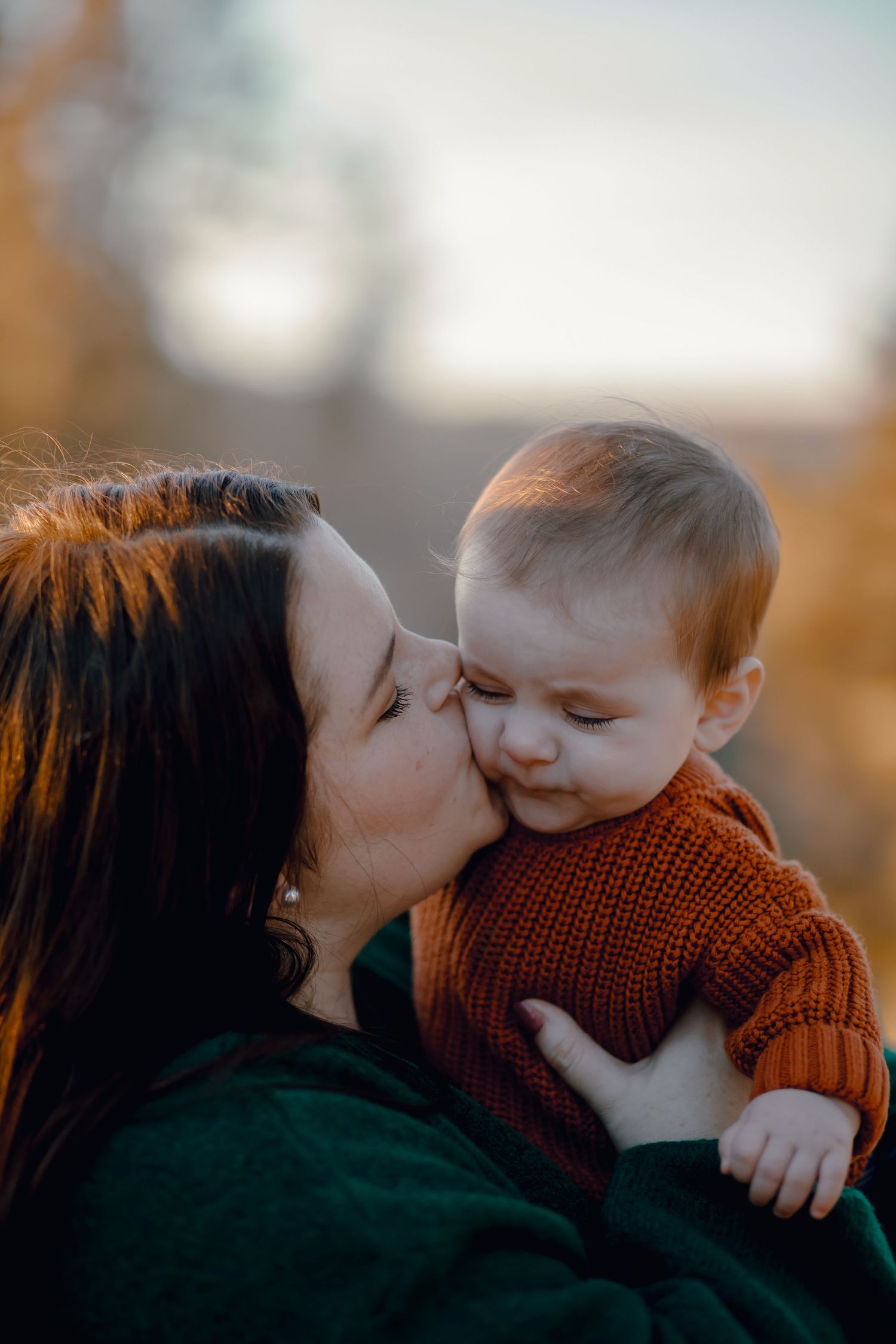 A tender moment captured showing a mother kissing her baby in a red sweater during a golden sunset.