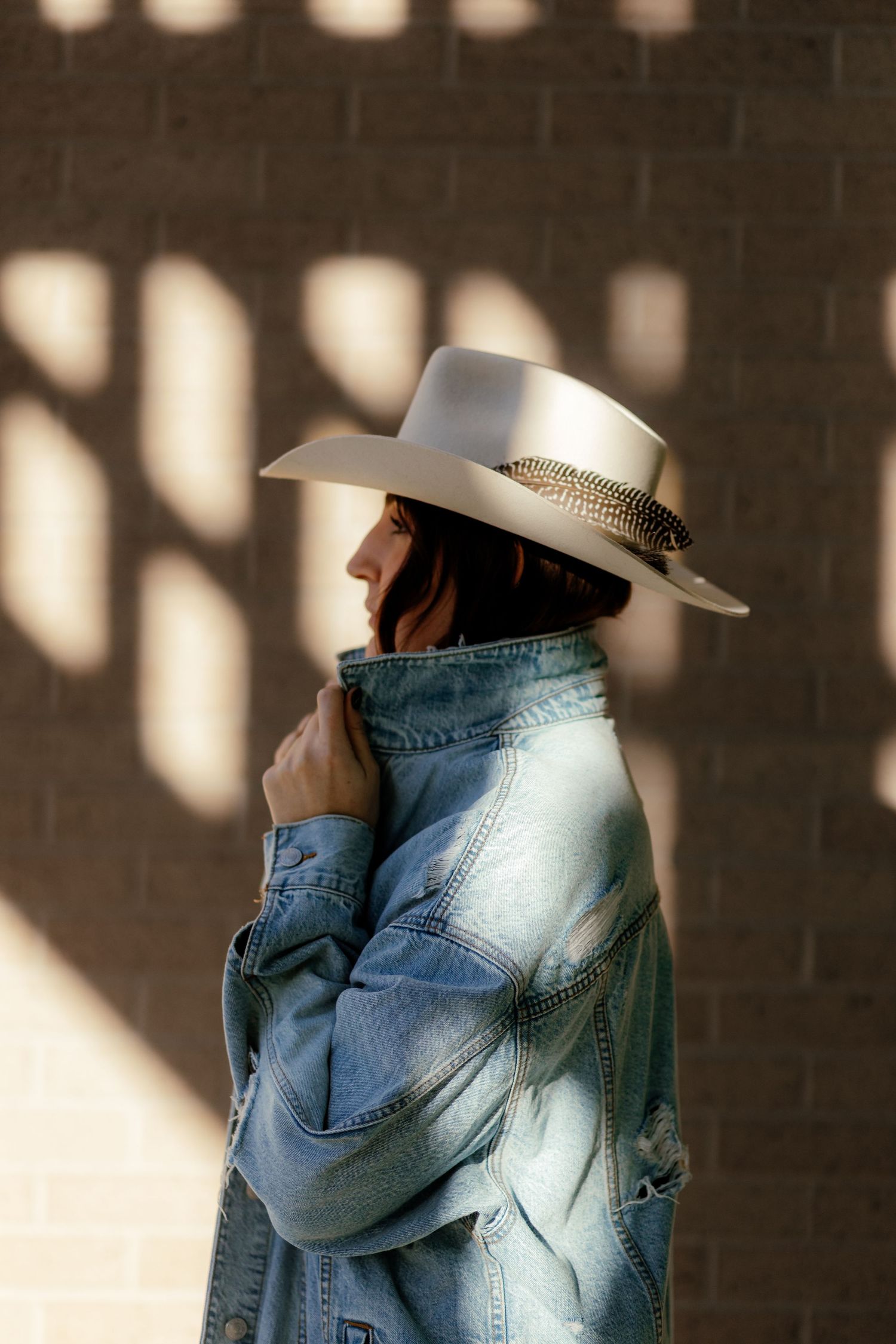 Side profile of figure wearing white cowboy hat and denim jacket stands against wall with dramatic window light shadows.