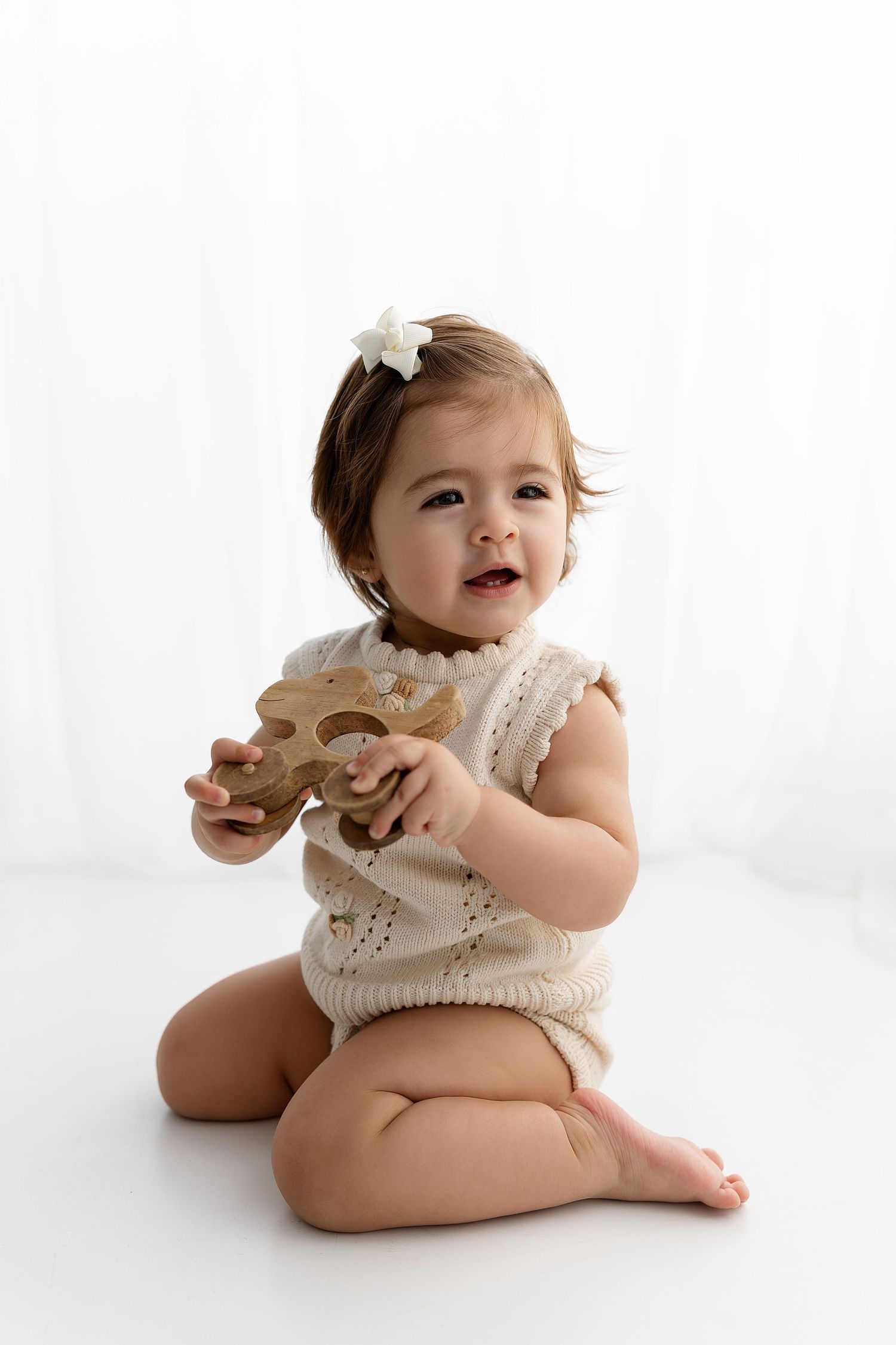 Happy toddler in cream outfit plays with wooden toy while sitting on white floor.