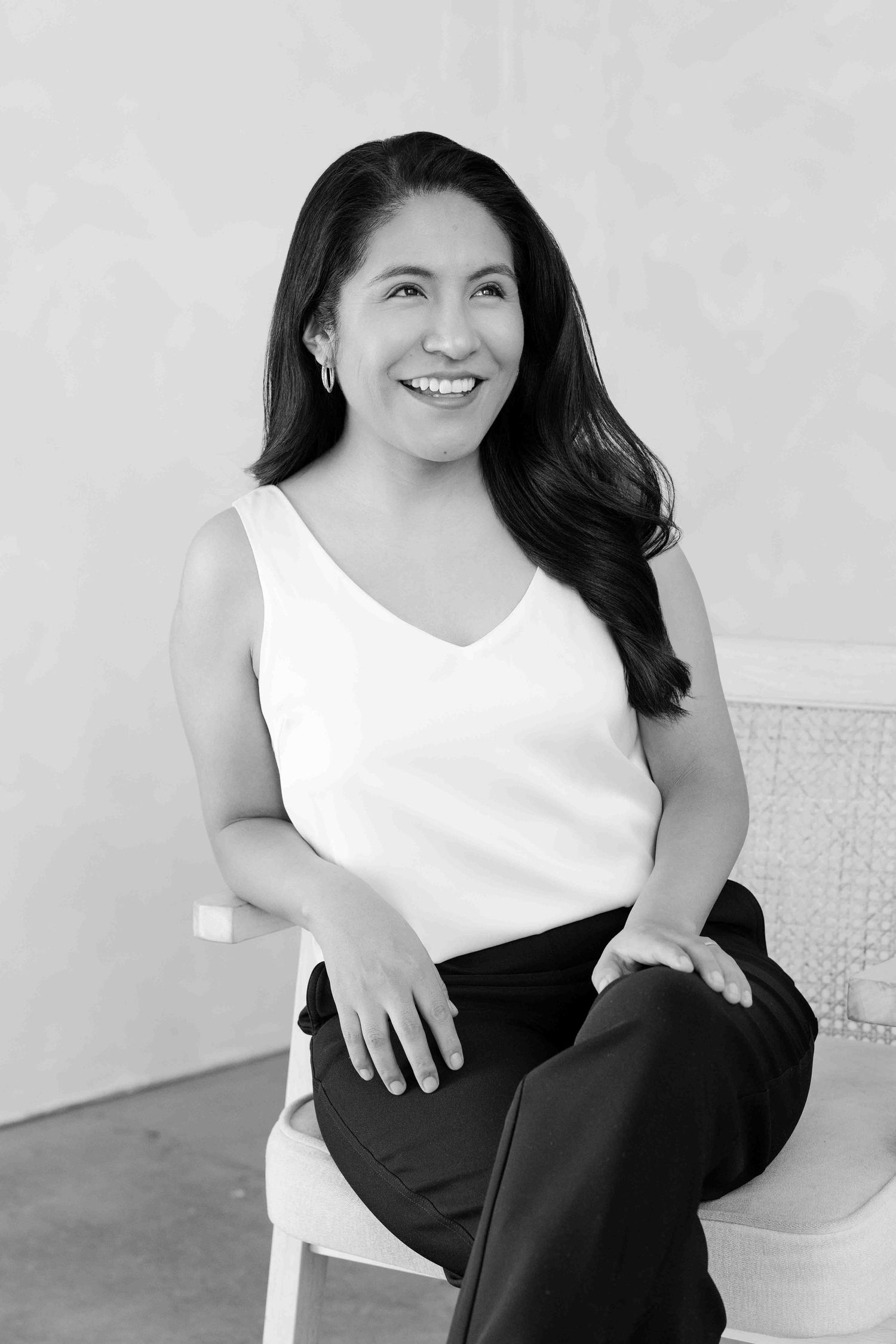 A black and white portrait of a woman in a white sleeveless top and dark pants sitting and smiling at the camera.