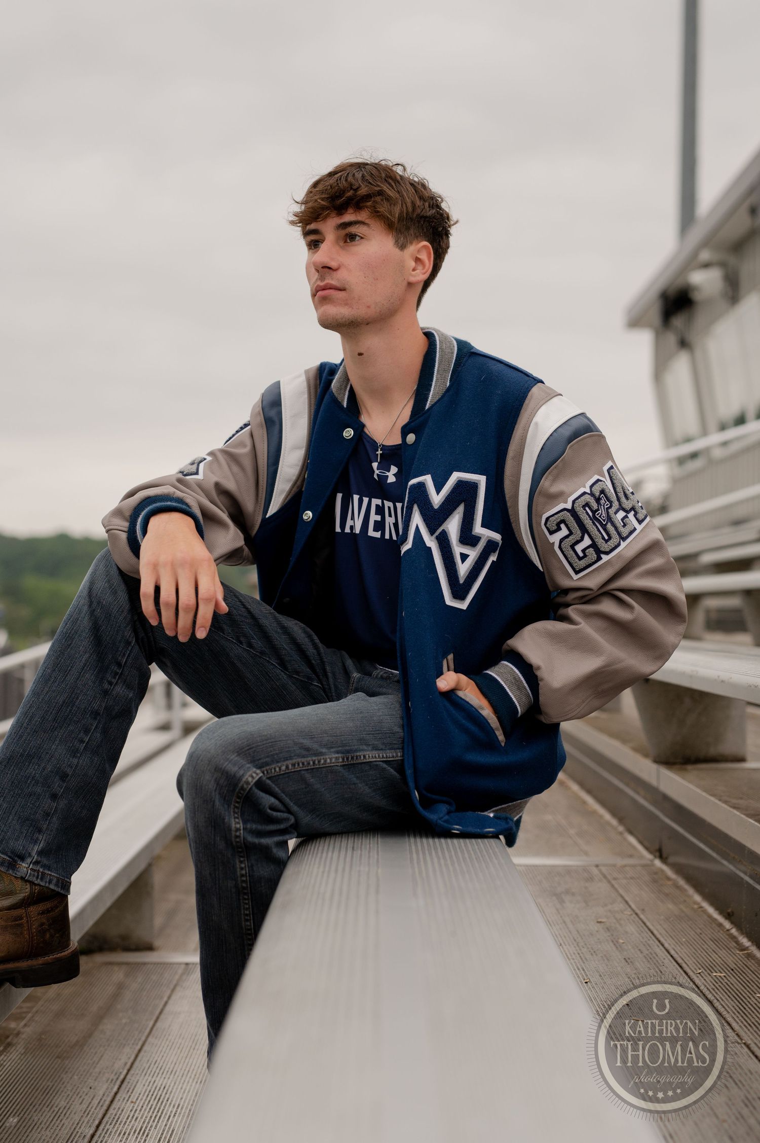 Student in blue and gray varsity letterman jacket poses thoughtfully on bleachers.