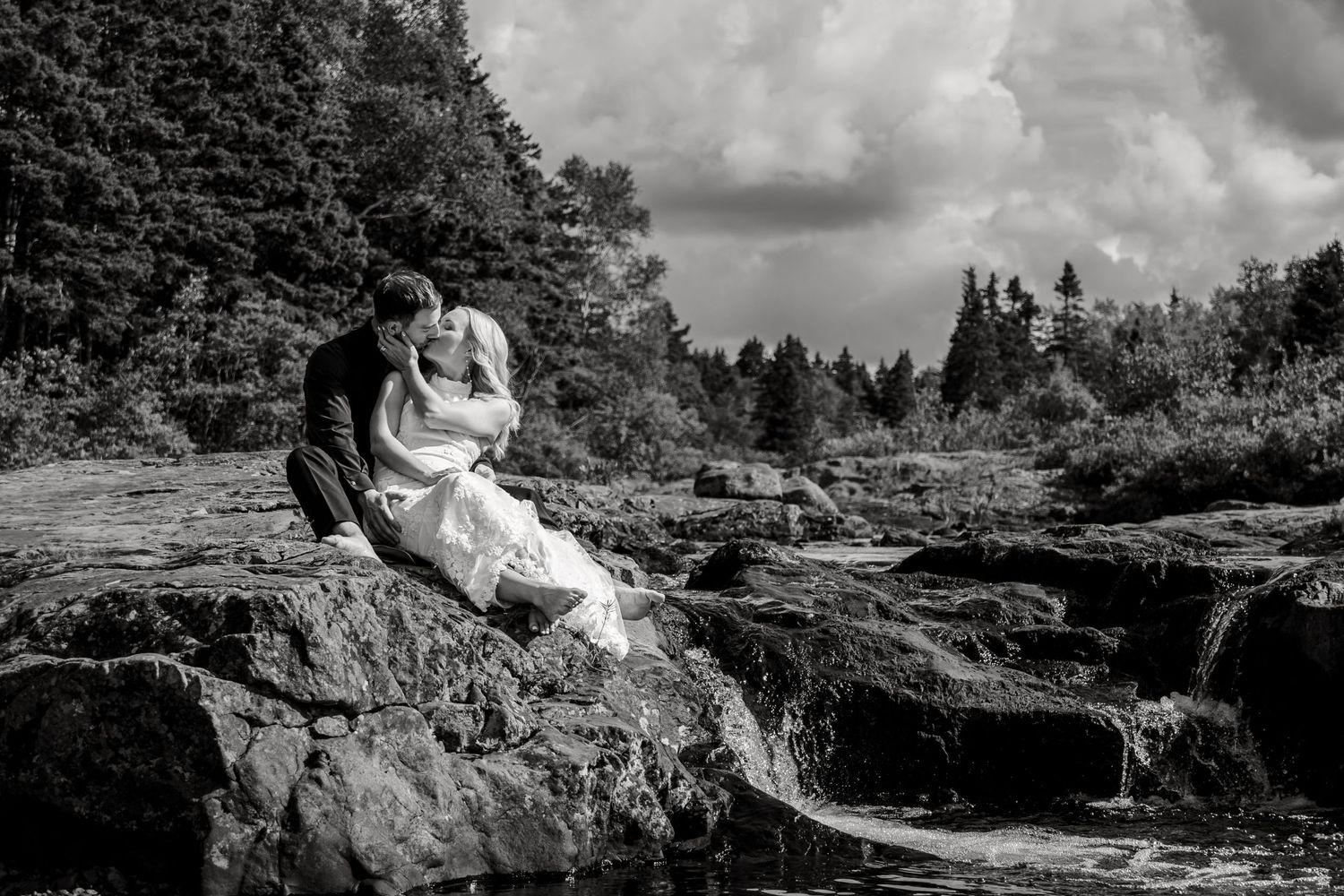 A couple sits together on rocky cliffs near a scenic waterfall in a dramatic black and white landscape scene.