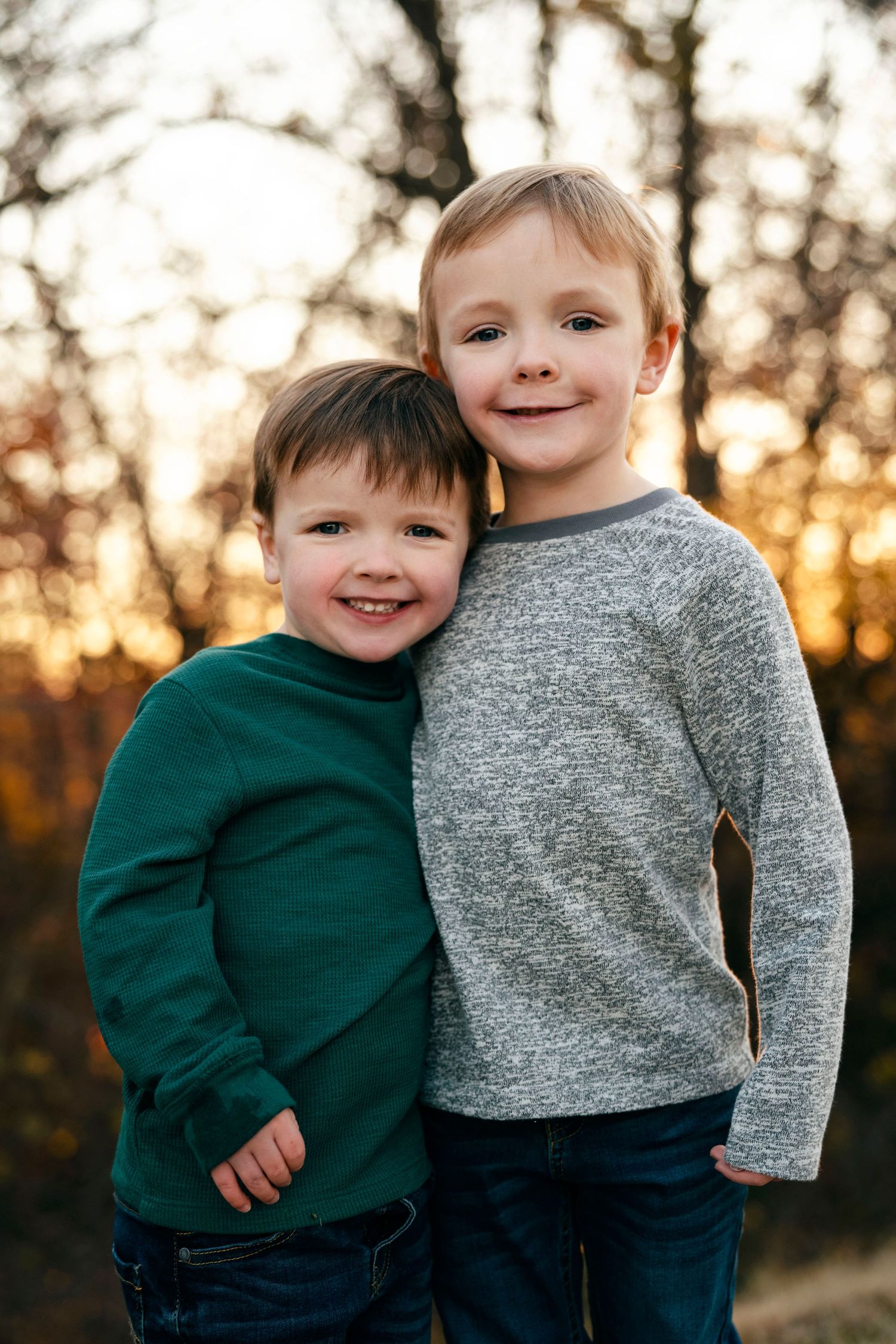 Two young boys in casual sweaters share a heartwarming embrace while smiling at sunset.