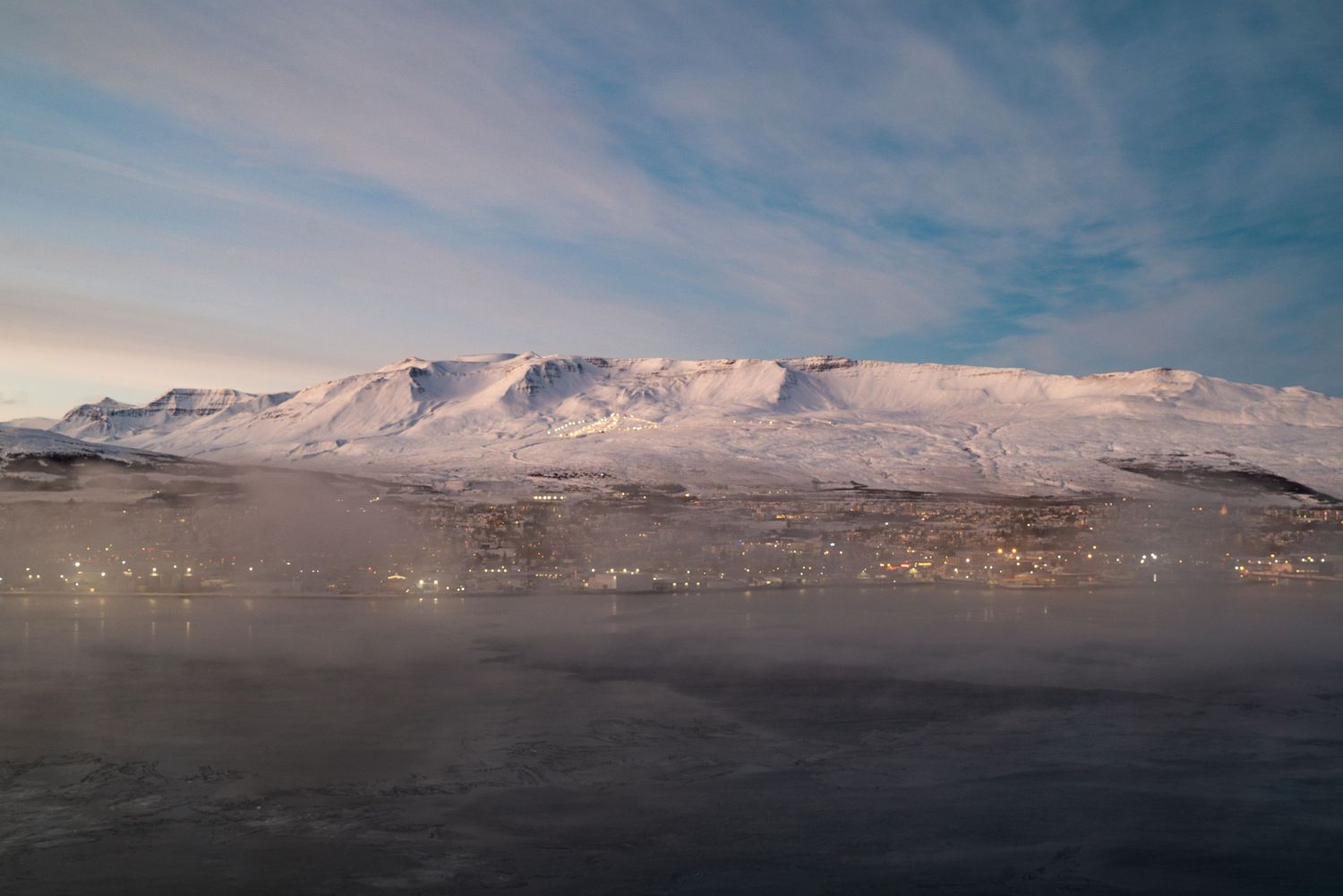 Snow-capped mountain range reflected in dark waters under a cloudy sky at dusk.