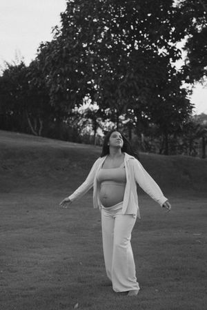 A pregnant woman in white clothing stands joyfully in a grassy field with trees in the background.