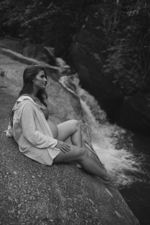 Black and white maternity portrait by waterfall showing woman in oversized shirt.