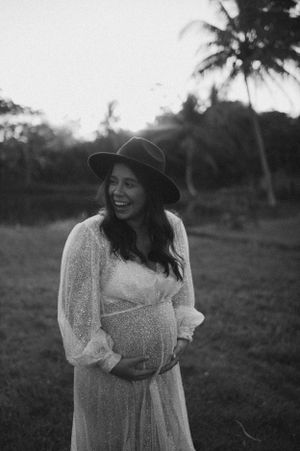 Expectant mother in white lace dress and hat standing in tropical setting at sunset.