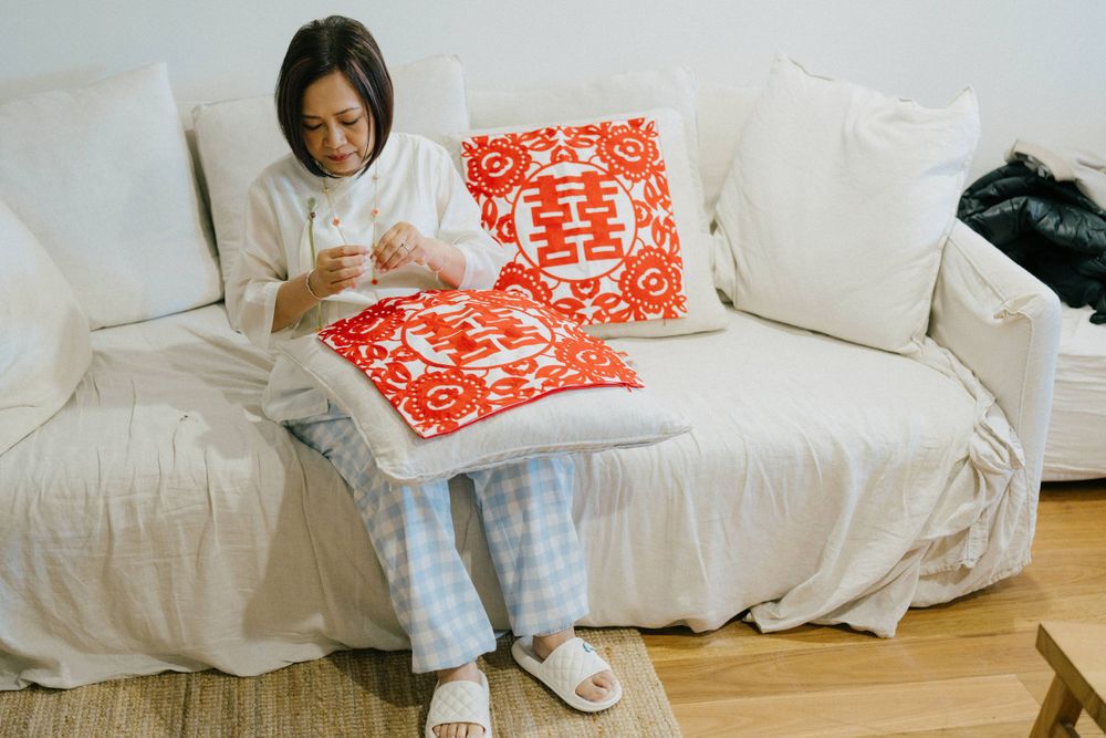 A person in plaid pajamas sits on a white couch with decorative red Chinese double happiness pillows.