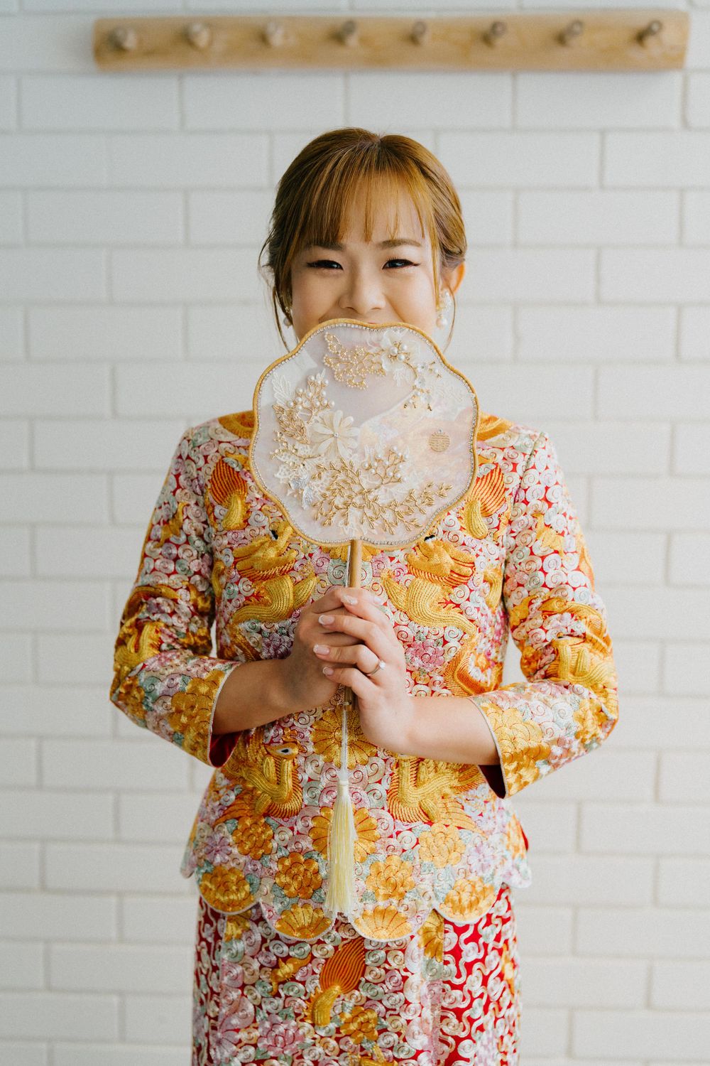Person in floral dress holding a decorative fan against white brick wall with wooden coat hooks.