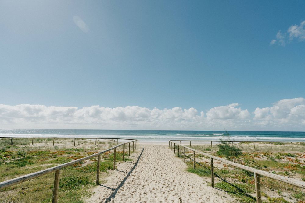 Sandy beach boardwalk path leading to ocean horizon under bright blue sky with white clouds.