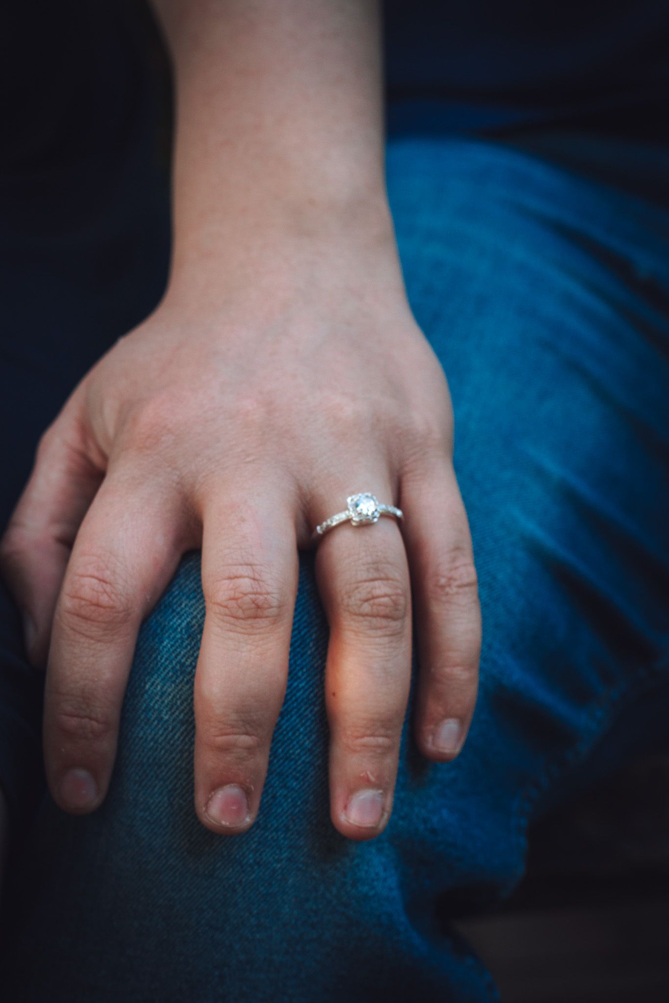 Close-up of an engagement ring on a hand resting against blue fabric.