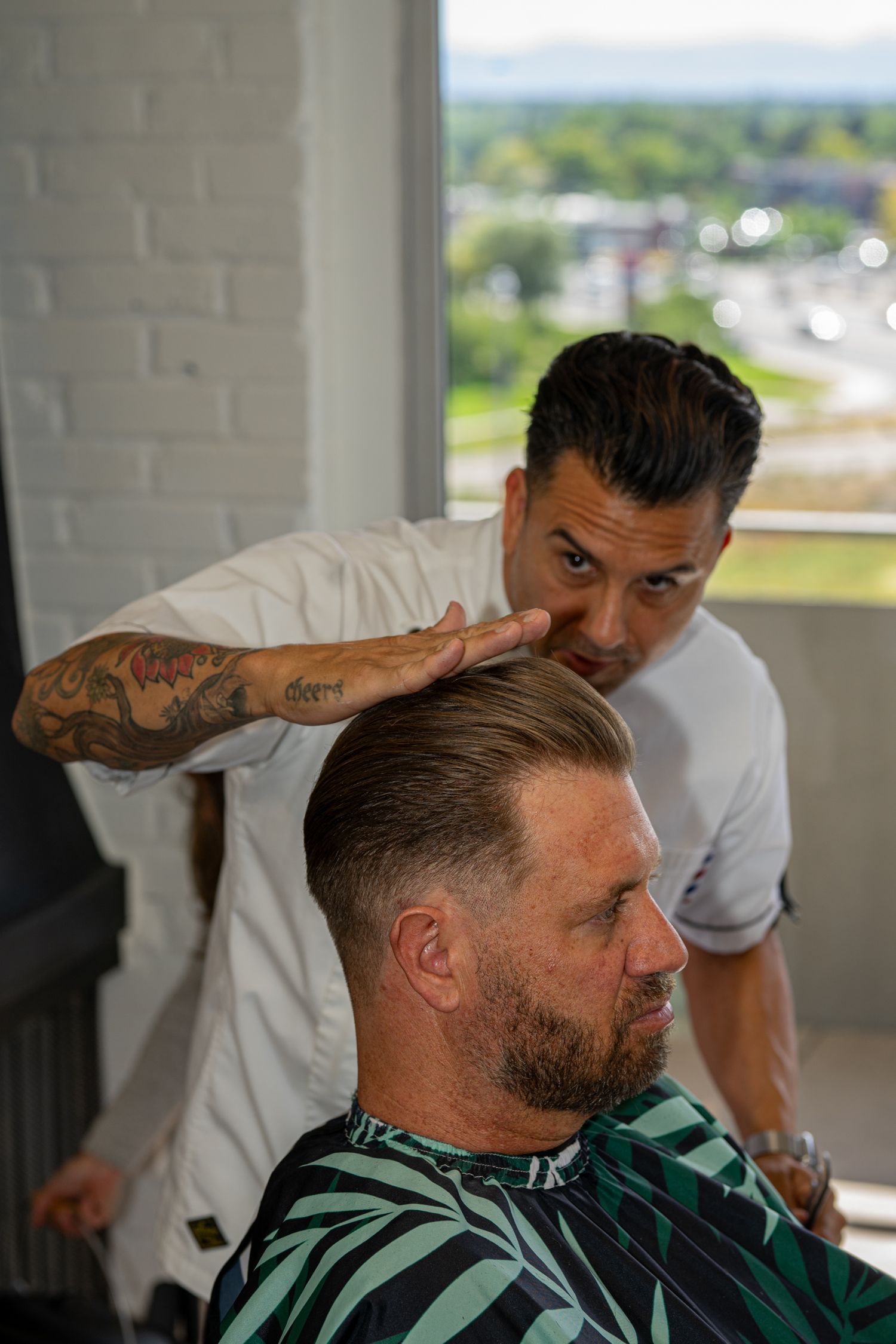 Tattooed barber styling client's hair in modern barbershop with city view.