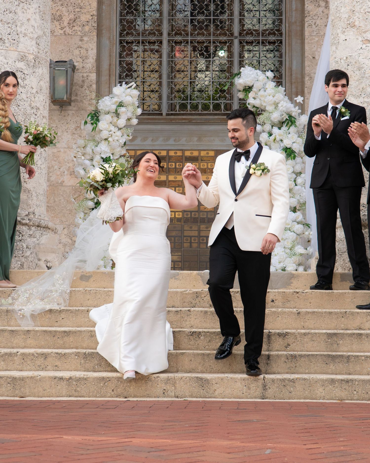 Bride and groom descend steps after wedding ceremony surrounded by floral arch and applauding guests.