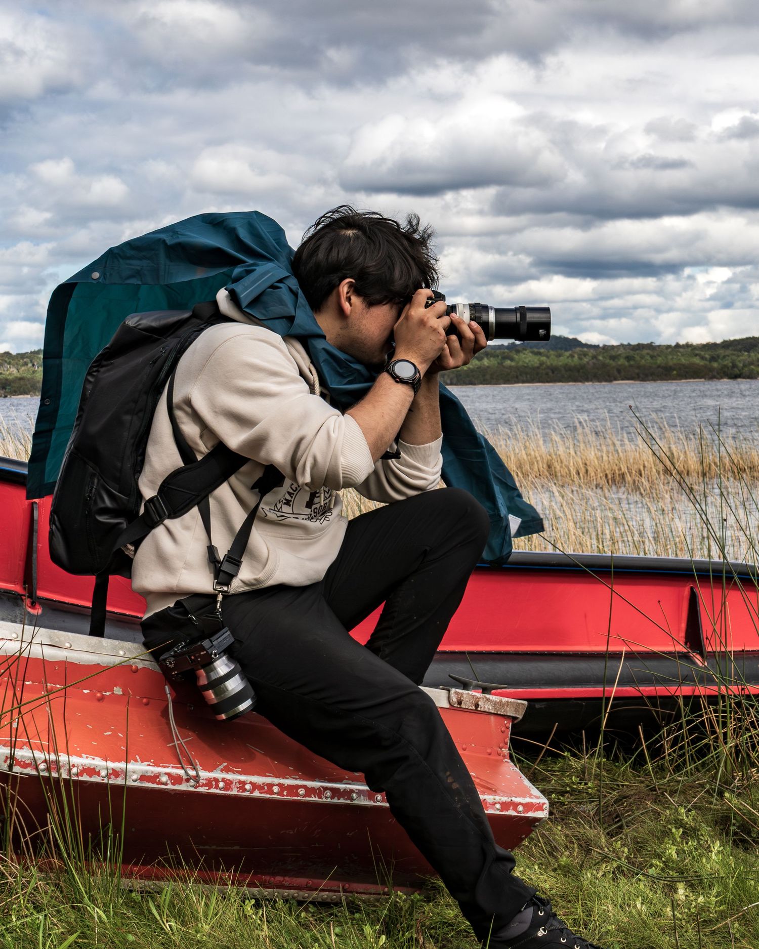 Fotógrafo con mochila tomando fotos desde canoa roja sobre cielo nublado y paisaje de humedal.