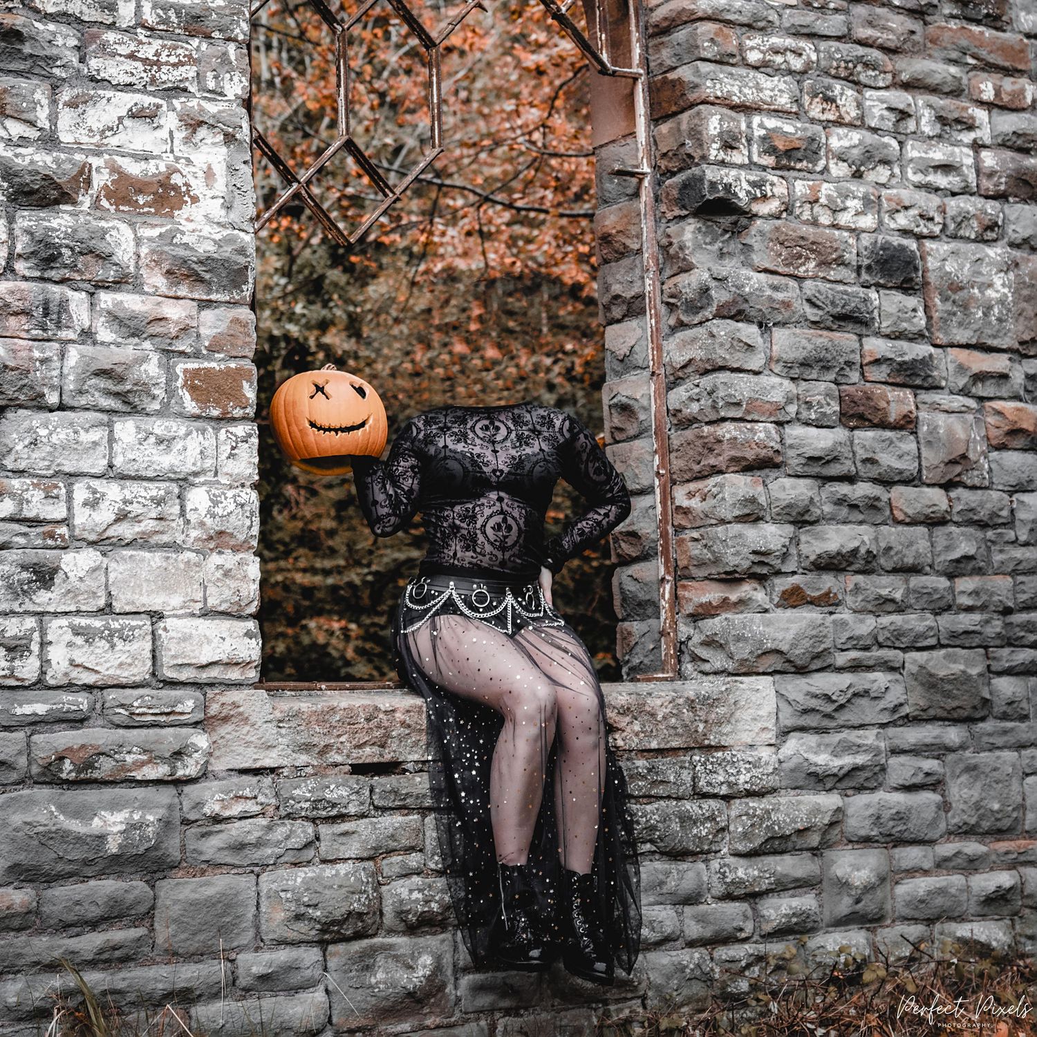 Halloween figure with pumpkin head in black lace dress sits on stone wall.