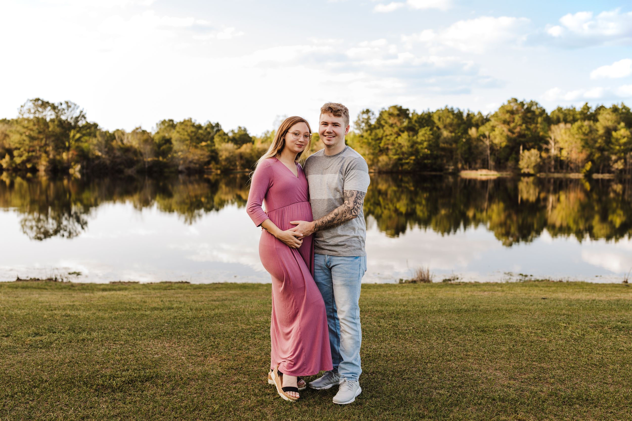 Couple embraces by serene lake at sunset with trees reflected in calm waters.