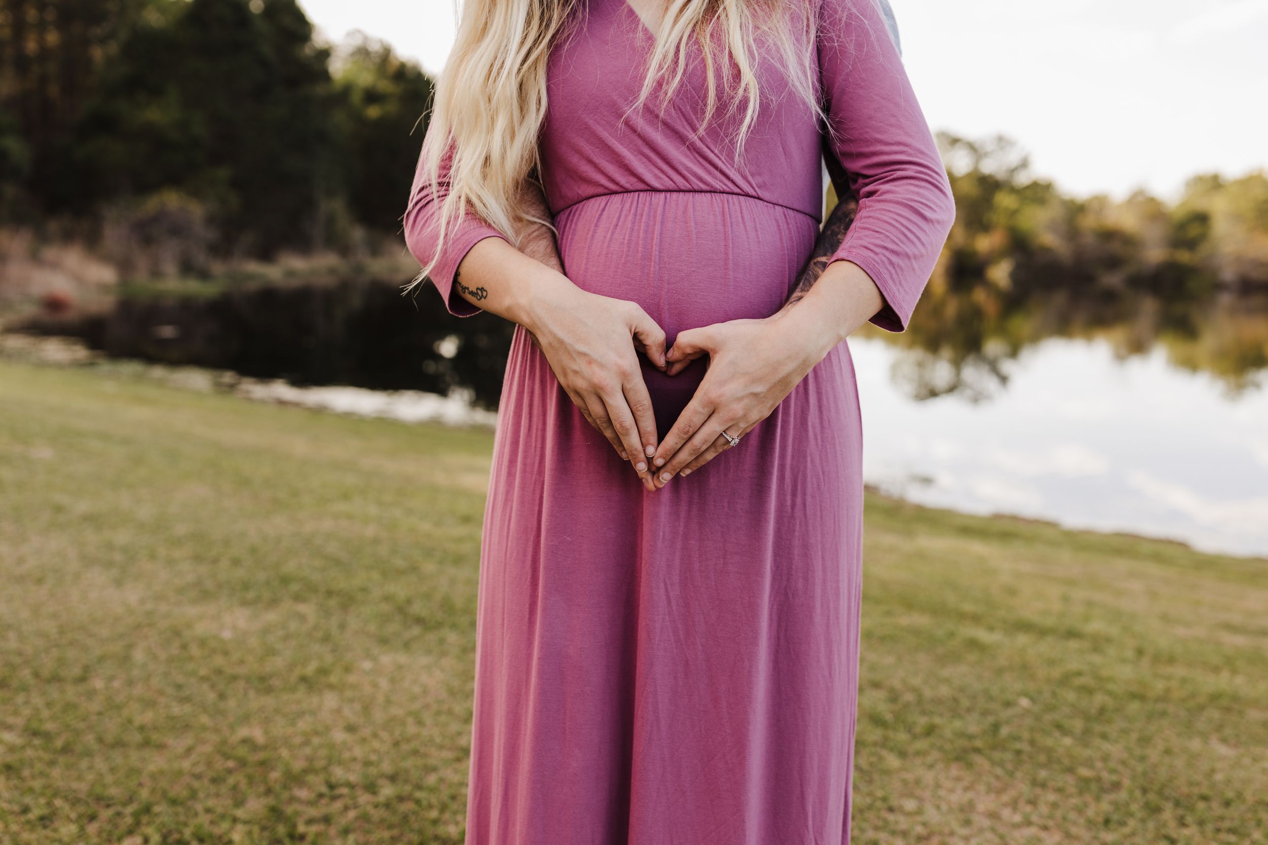 A maternity photo showing a woman in a flowing pink dress making a heart shape with her hands.