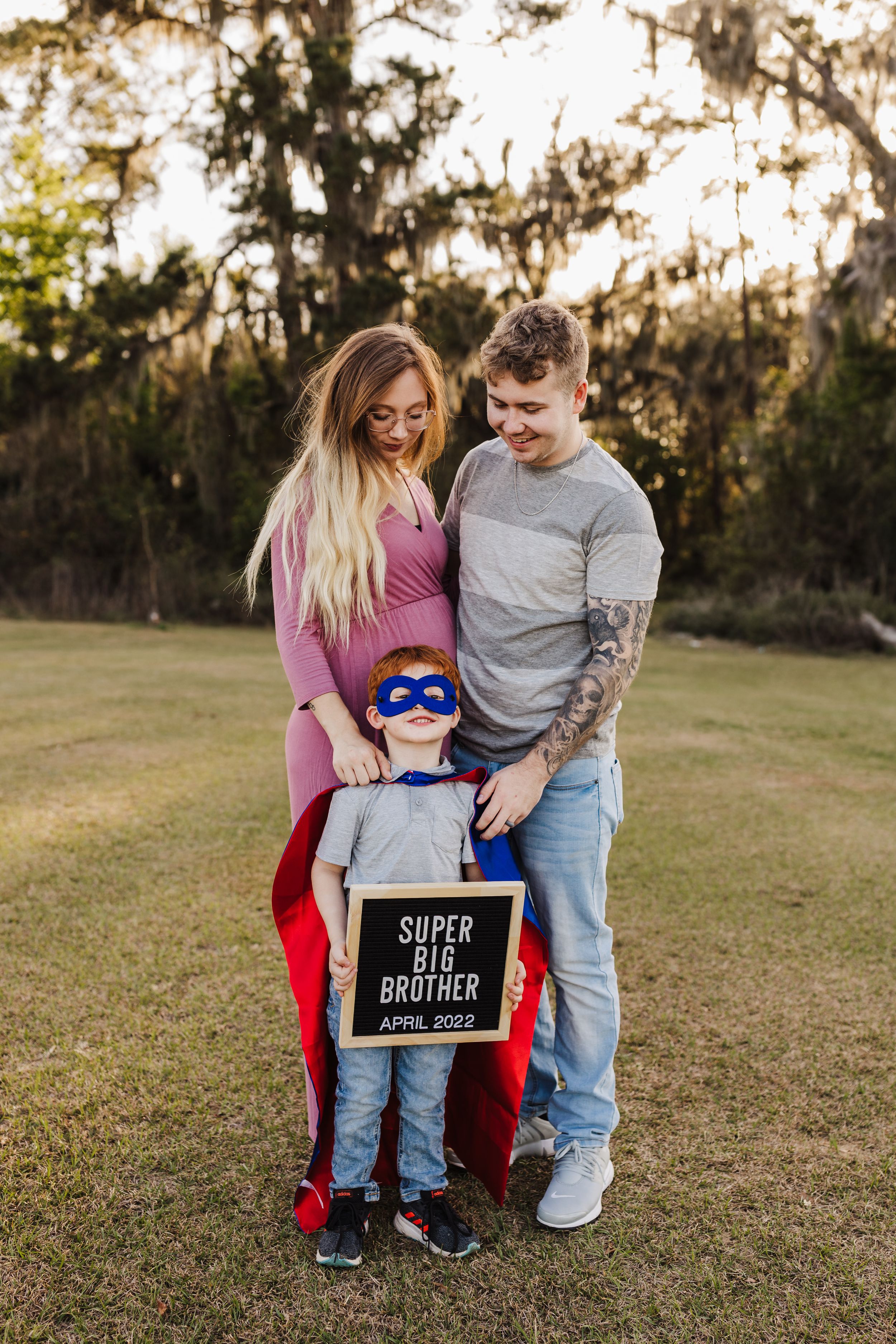 A family pregnancy announcement featuring a child in superhero costume with a letter board.