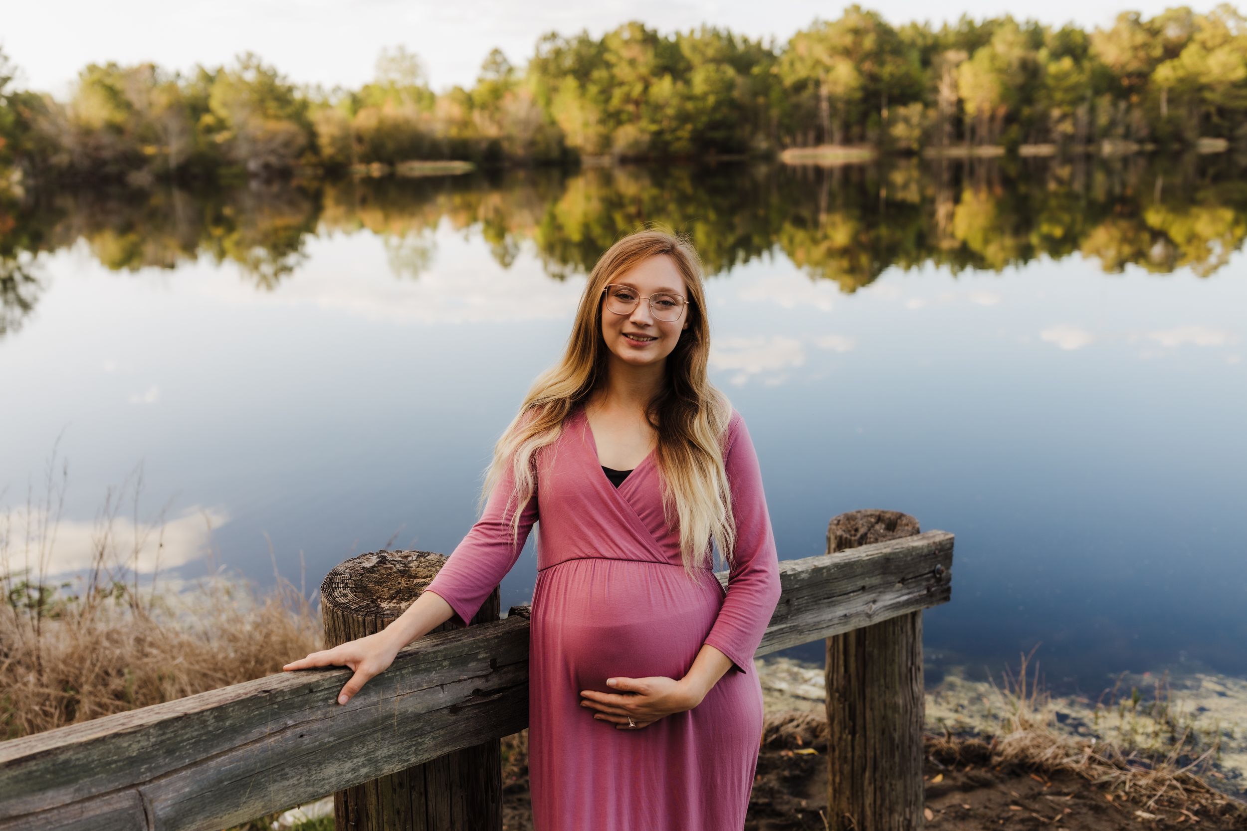 An expectant mother in a pink maxi dress stands on a wooden dock by a serene lake.