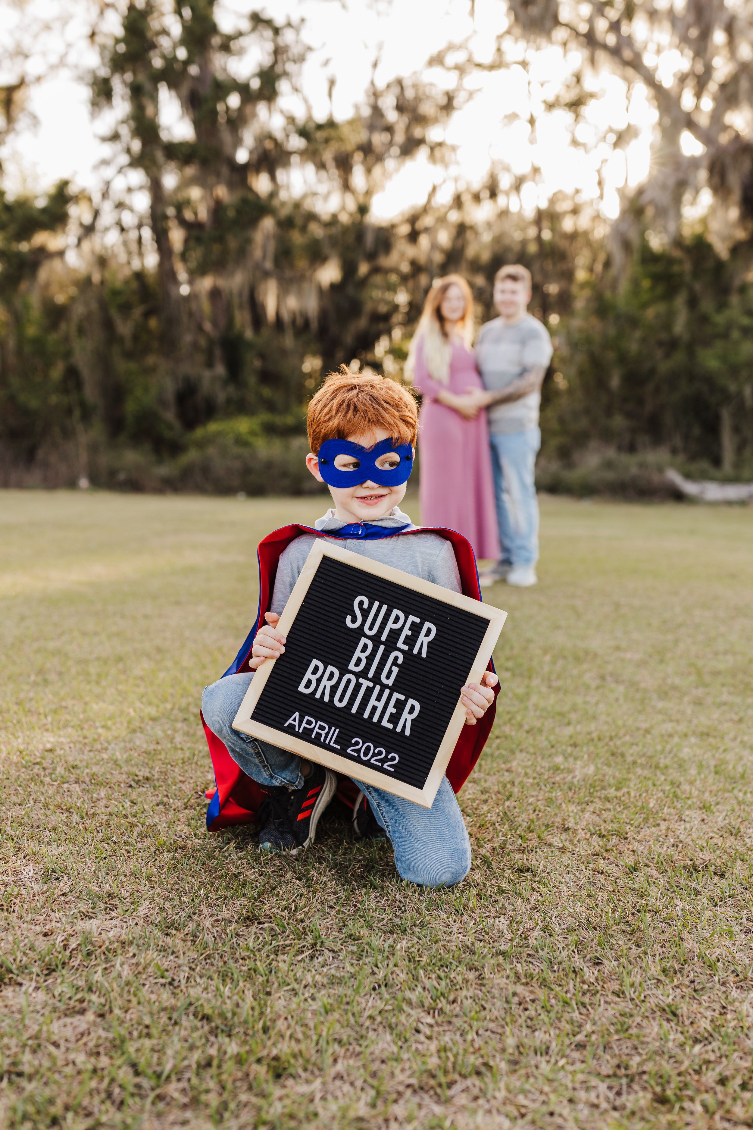 A creative pregnancy announcement with a child dressed as superhero in the foreground.