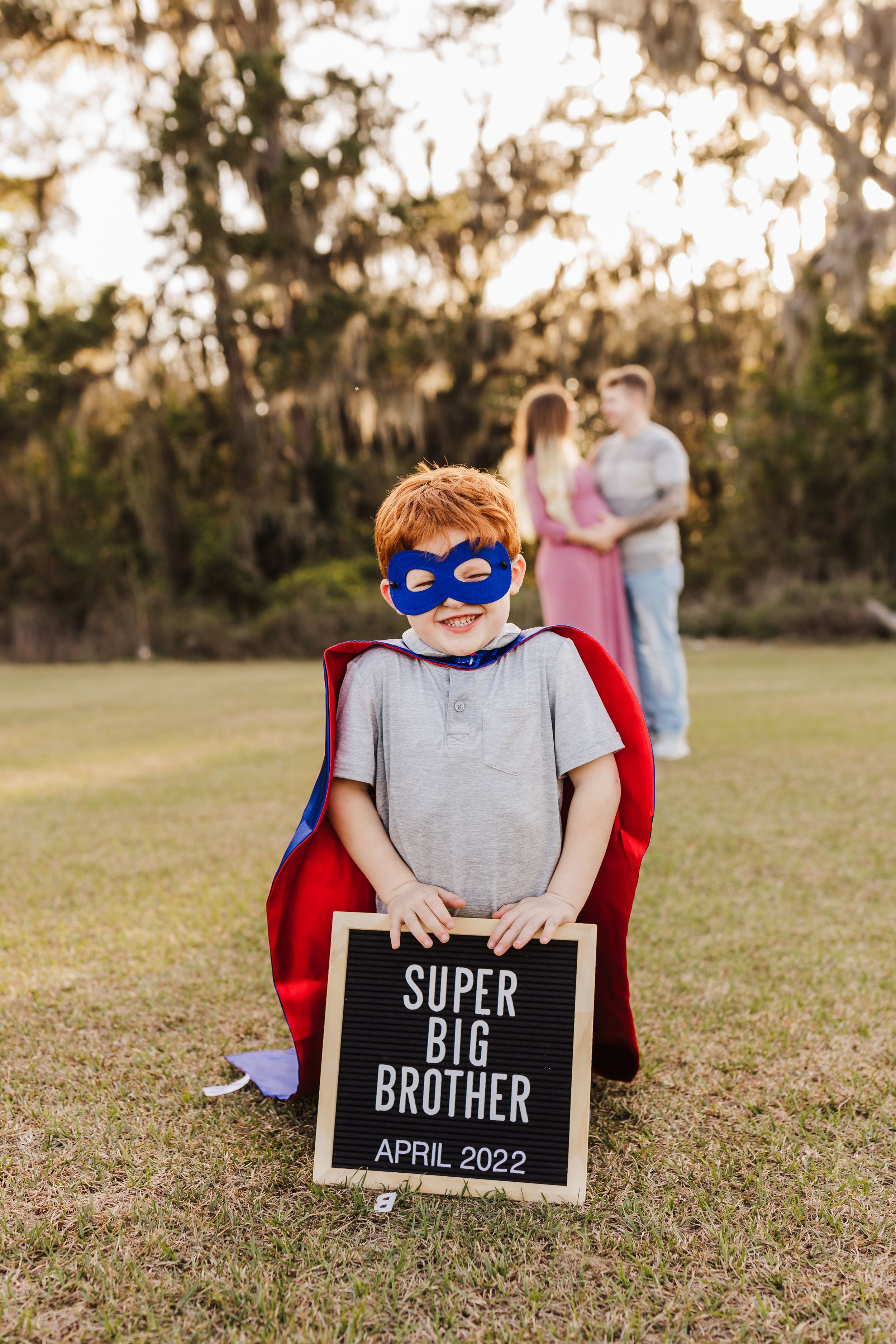 A child in superhero costume holds a letter board announcing upcoming big brother status.