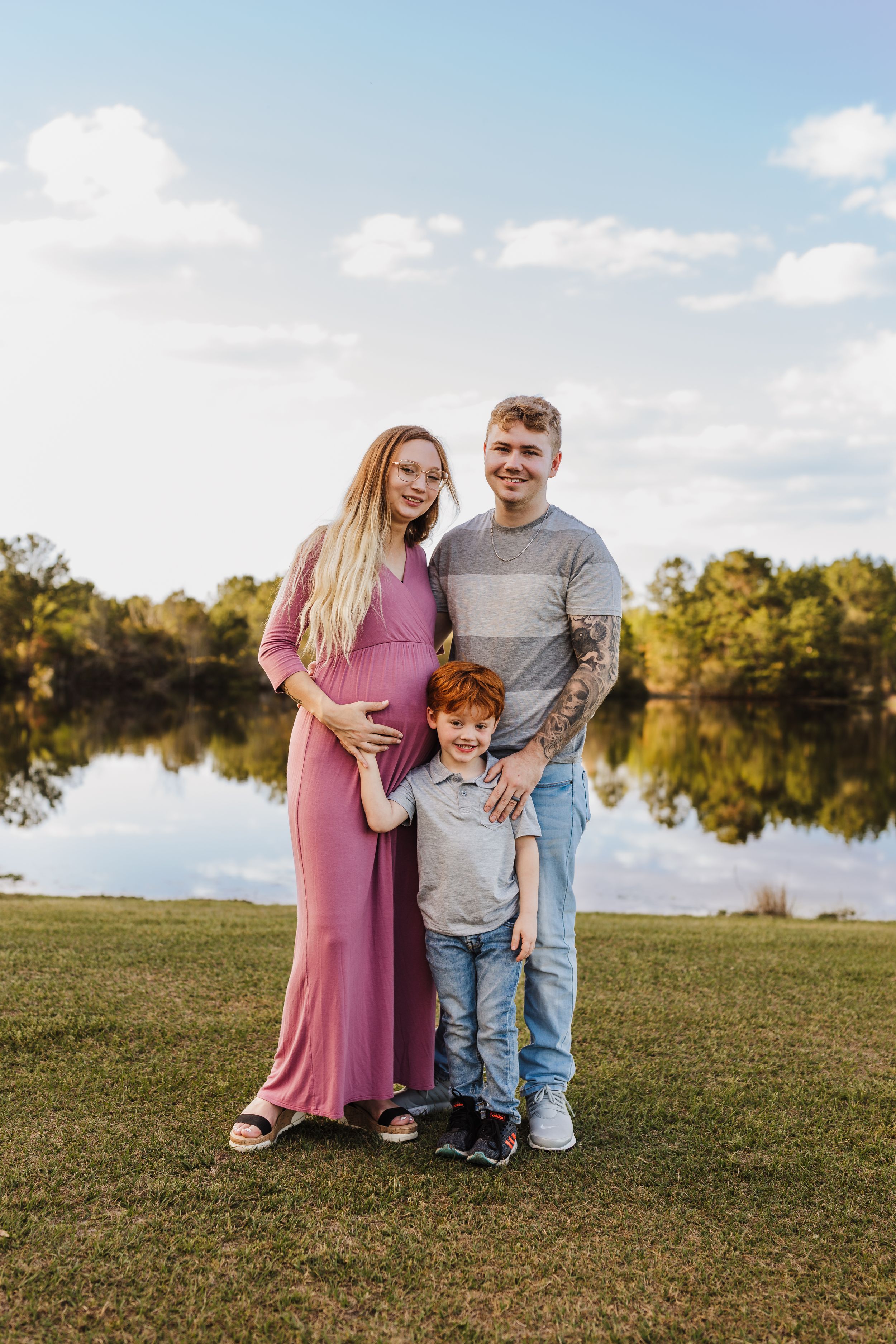 A family portrait by a lake with expectant mother in pink dress and scenic background.
