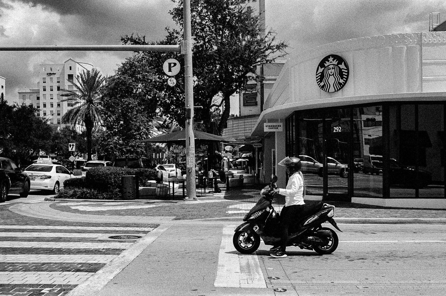 Black and white street scene showing a scooter at a crosswalk near a Starbucks cafe storefront.