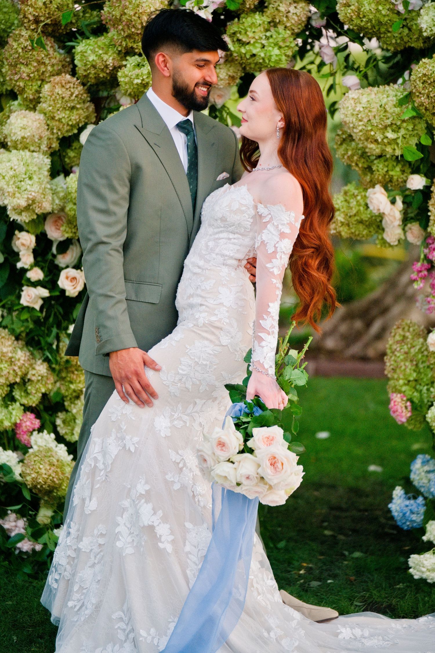 A bride in a lacy white dress and groom in a sage green suit embrace in a garden surrounded by hydrangeas and roses.