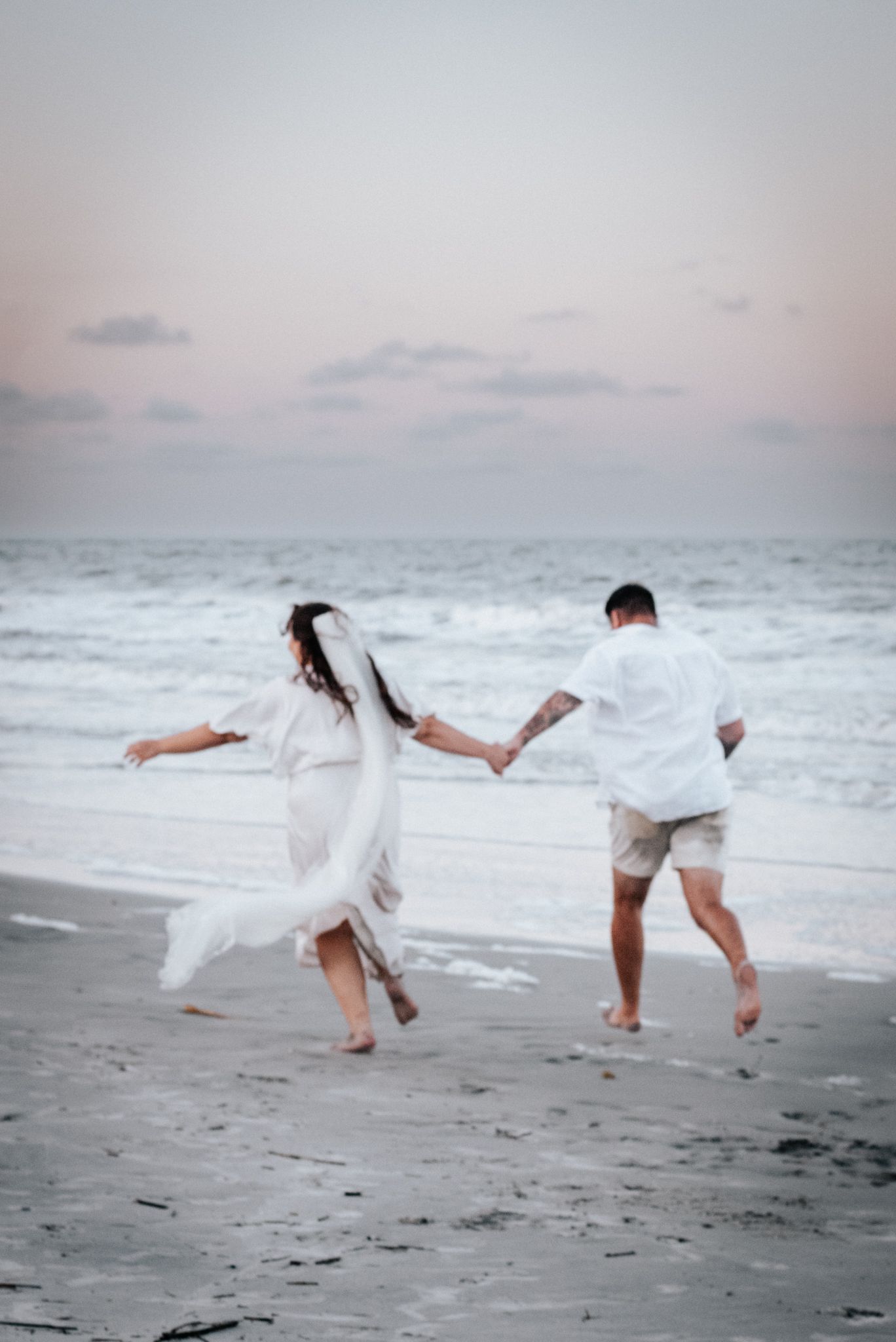 Couple in white clothing playfully running and splashing along a misty beach shoreline.