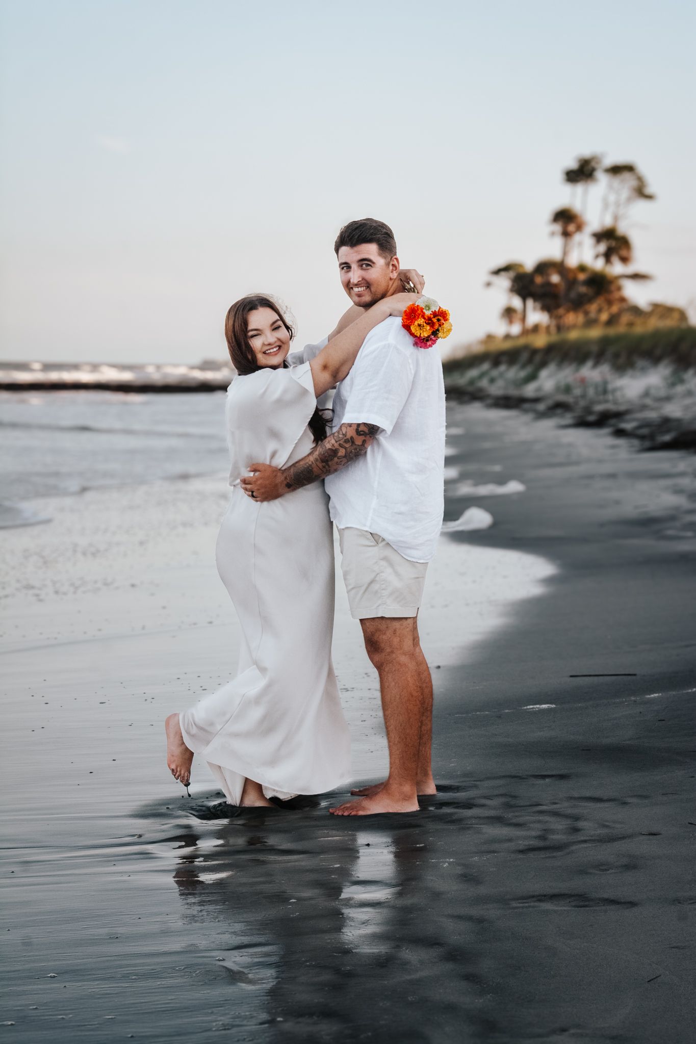 Couple in white attire embracing on beach at sunset.