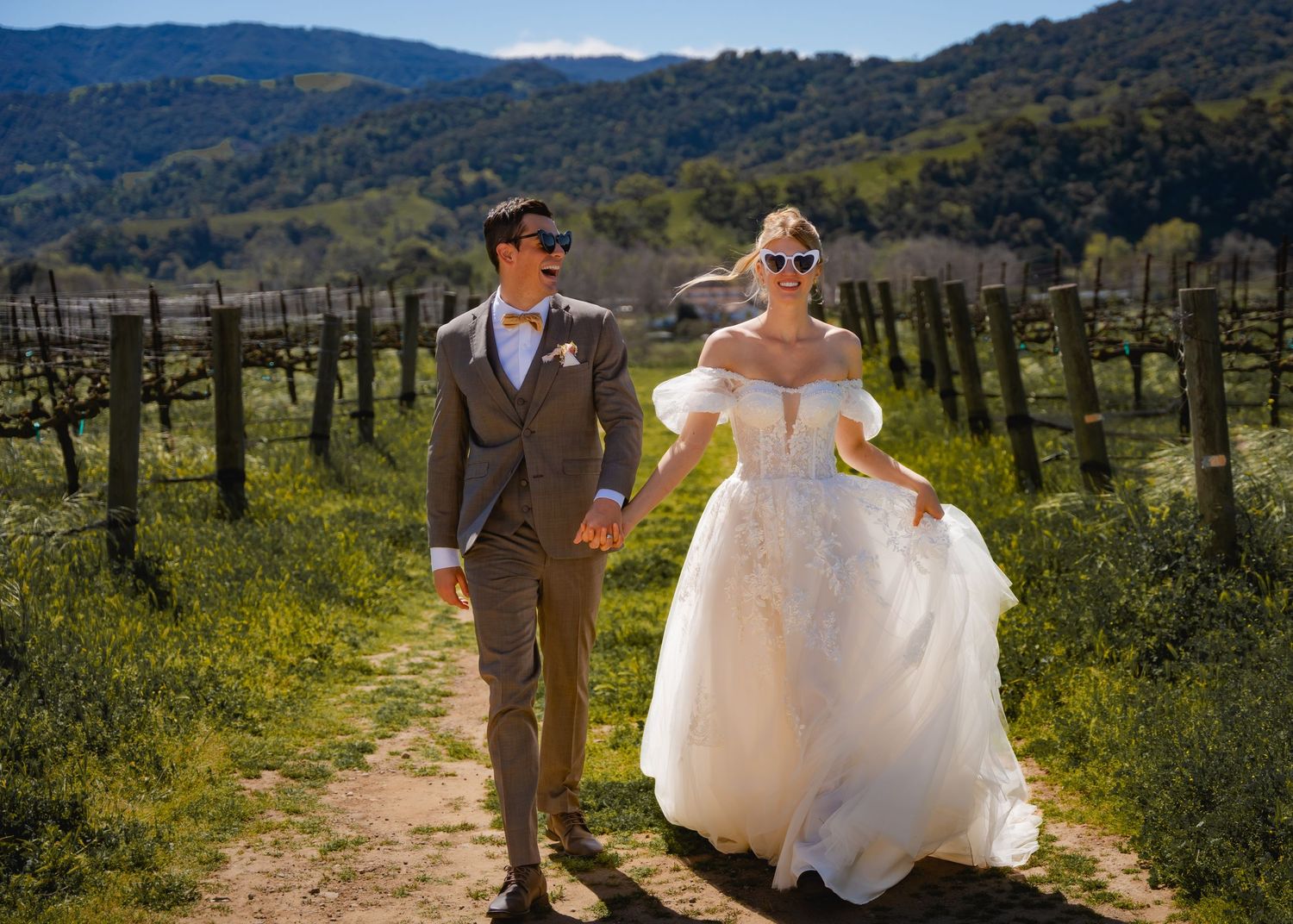 A couple in wedding attire walks through a sunny vineyard with rolling hills in the background.