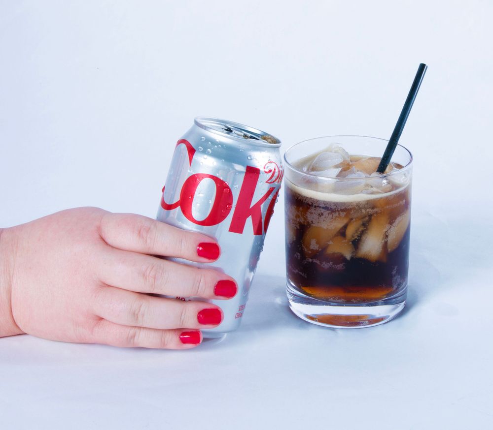 Hand with red nail polish holds Diet Coke can next to glass of iced soda with straw.