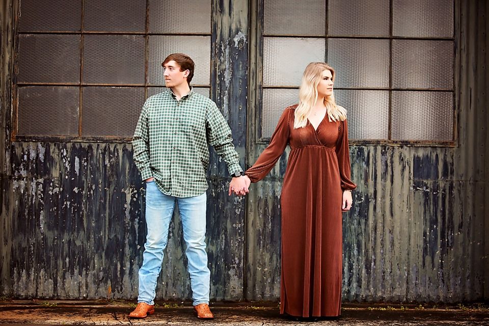 Couple holding hands in contrasting outfits against industrial wall background.