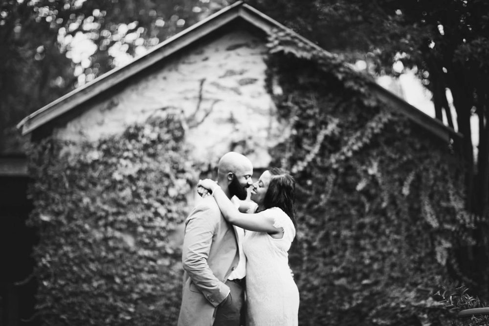 A black and white photograph of a couple embracing near an ivy-covered building.