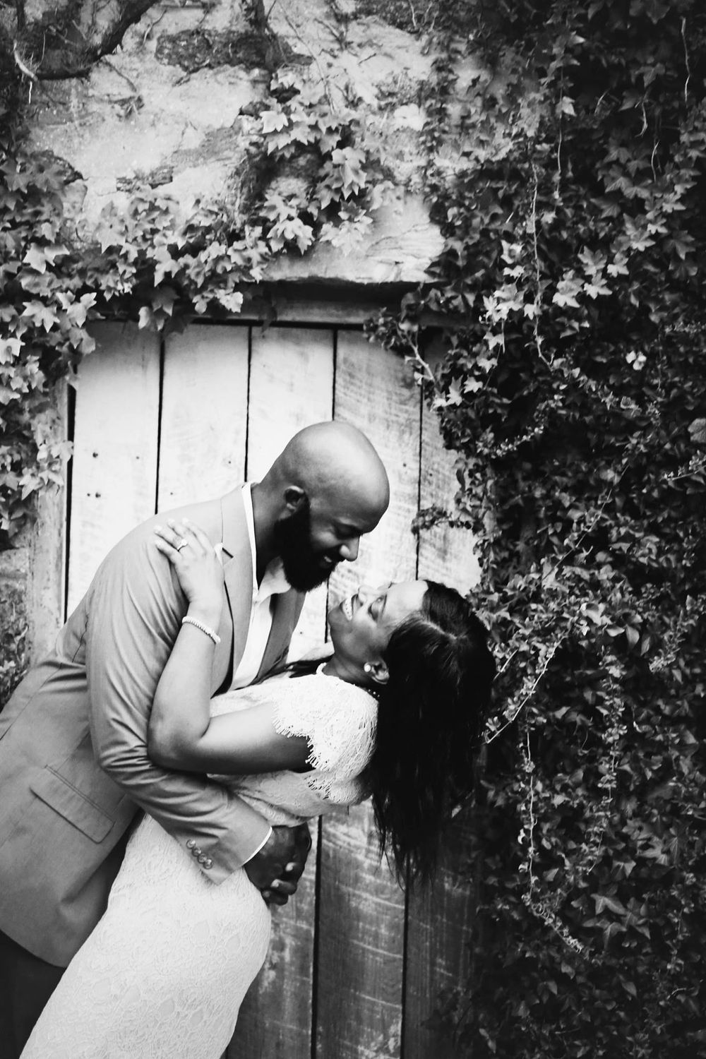 A romantic black and white photo of a couple embracing against an ivy-covered wall.