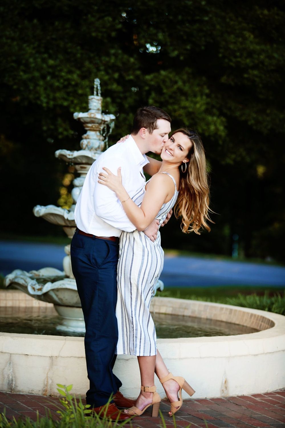 A couple embraces by an ornate fountain in a manicured garden setting.
