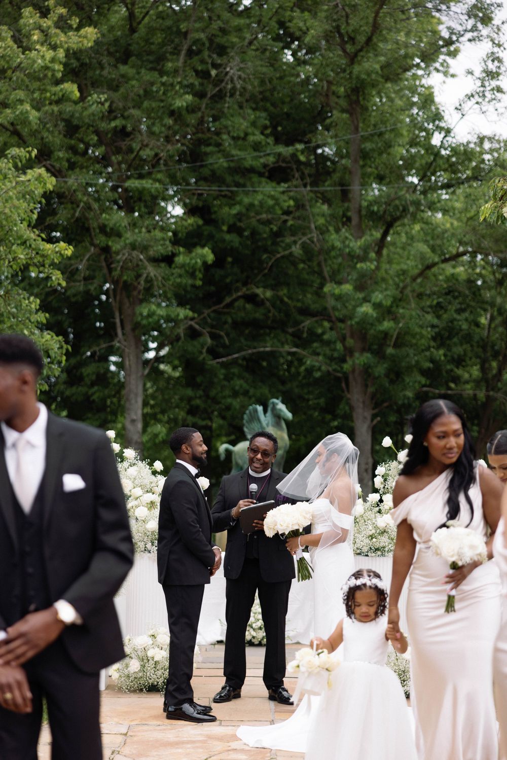 Wedding party gathers on stone pathway surrounded by lush green trees and white flowers.
