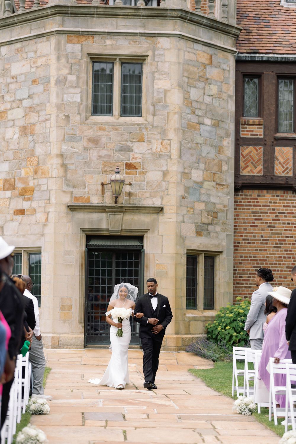 Wedding ceremony setup outside stone building with white chairs and floral decorations.