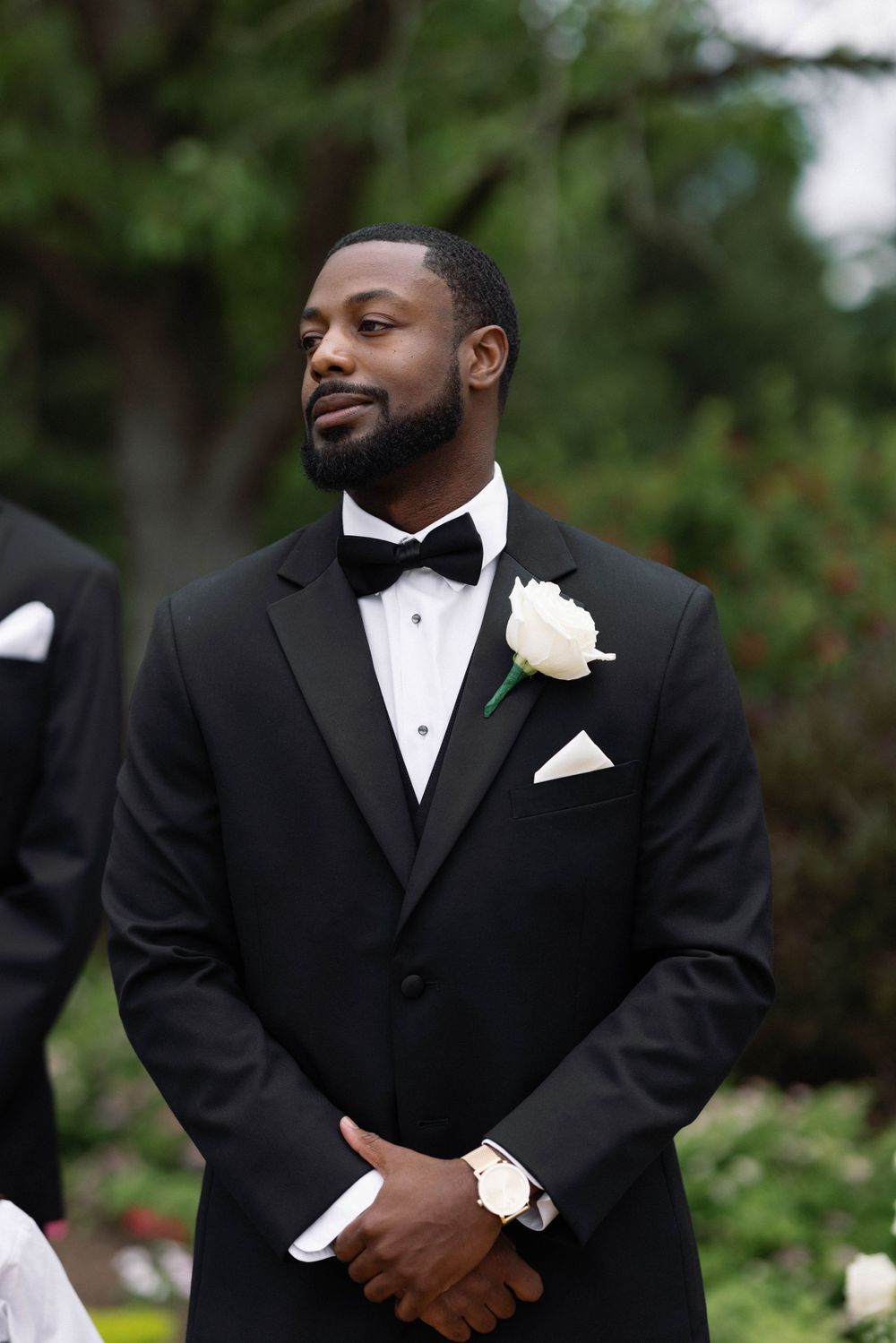 Groom in classic black tuxedo with white boutonniere smiles during outdoor wedding ceremony.