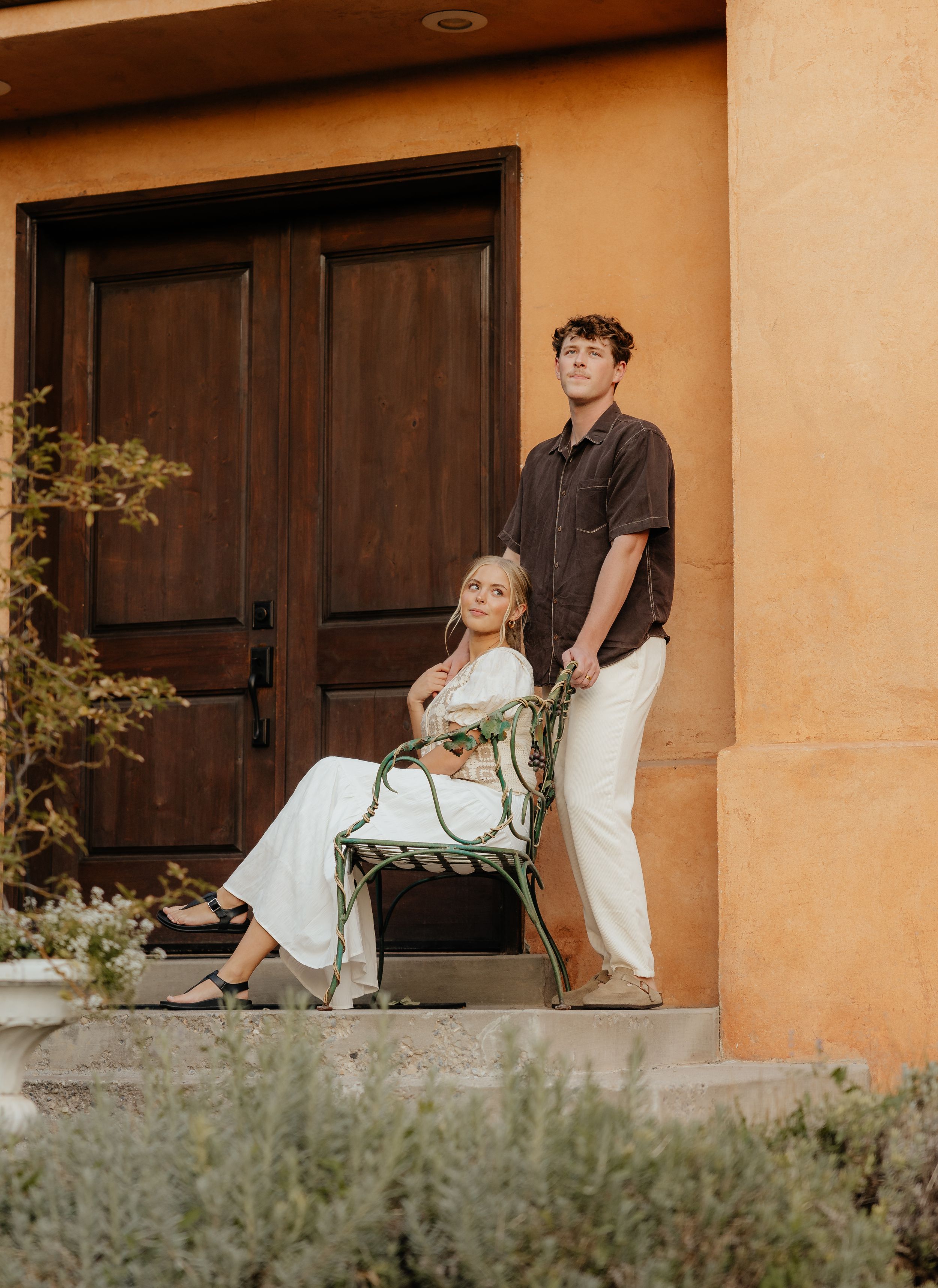 An engaged couple poses elegantly outside a warm adobe building with a dark wood door at golden hour.