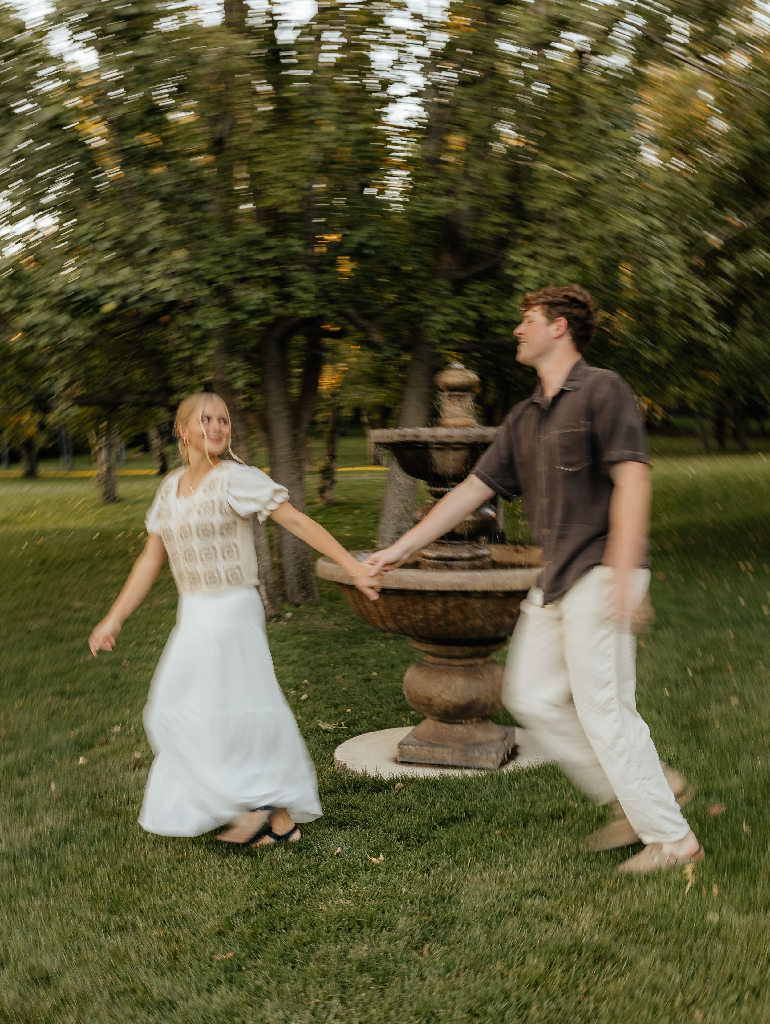 A couple playfully dances and spins holding hands near a stone fountain in a lush green park.